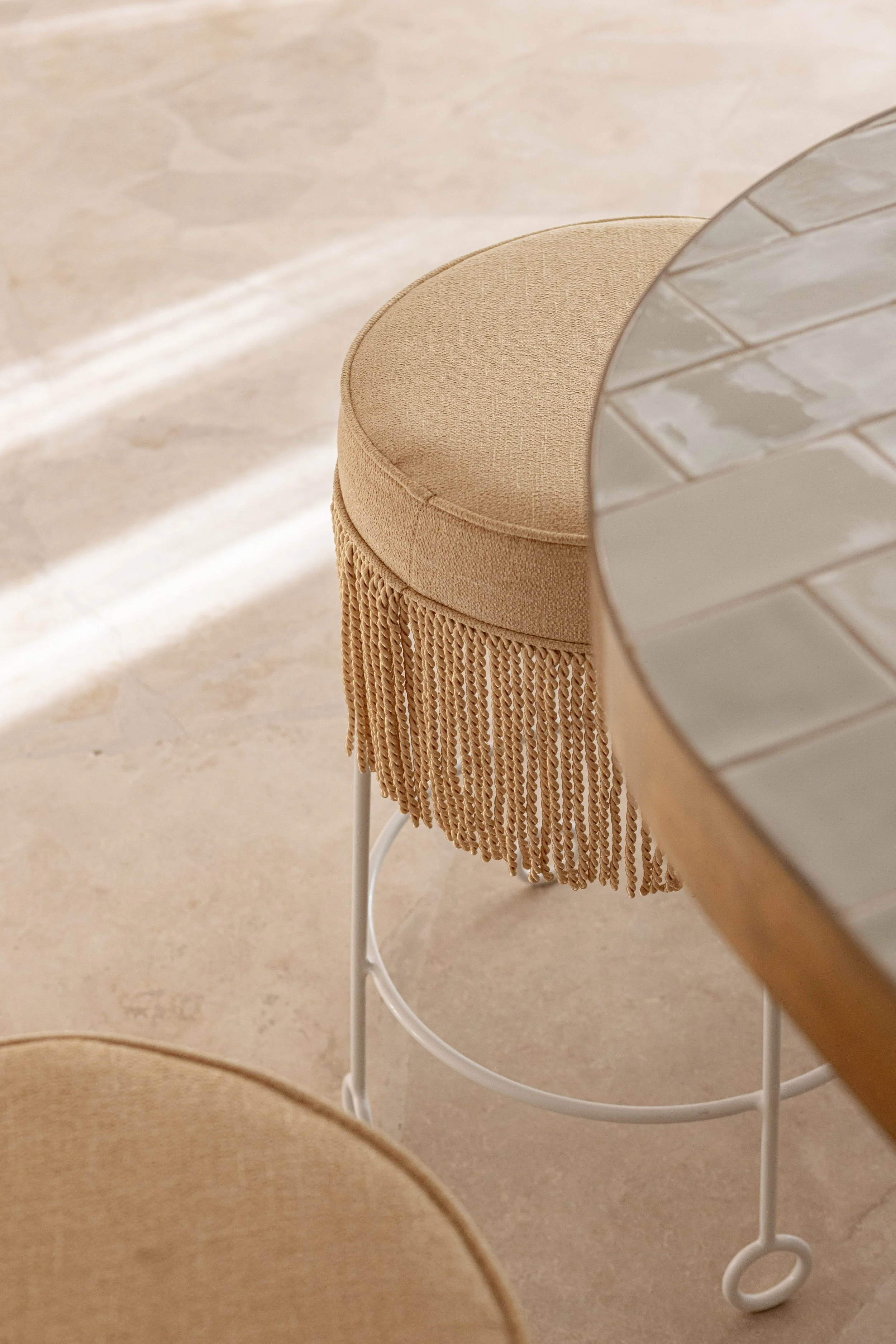 Beige upholstered barstool with fringe detail next to a tiled countertop.