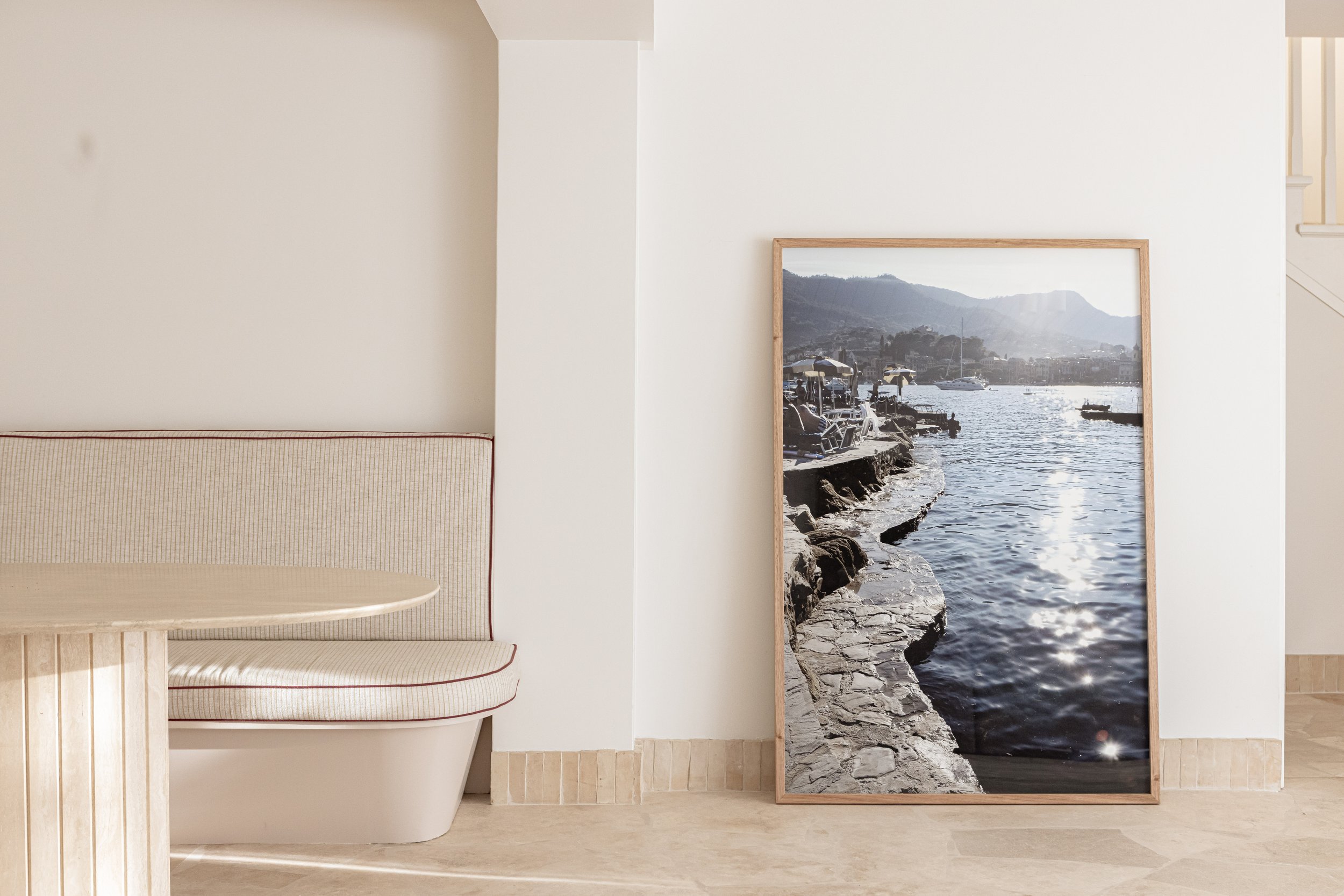 A framed photo of a rocky shoreline with boats on the water and mountains in the background leaning against a white wall inside a room with beige floors and a built-in corner bench with light-colored upholstery.