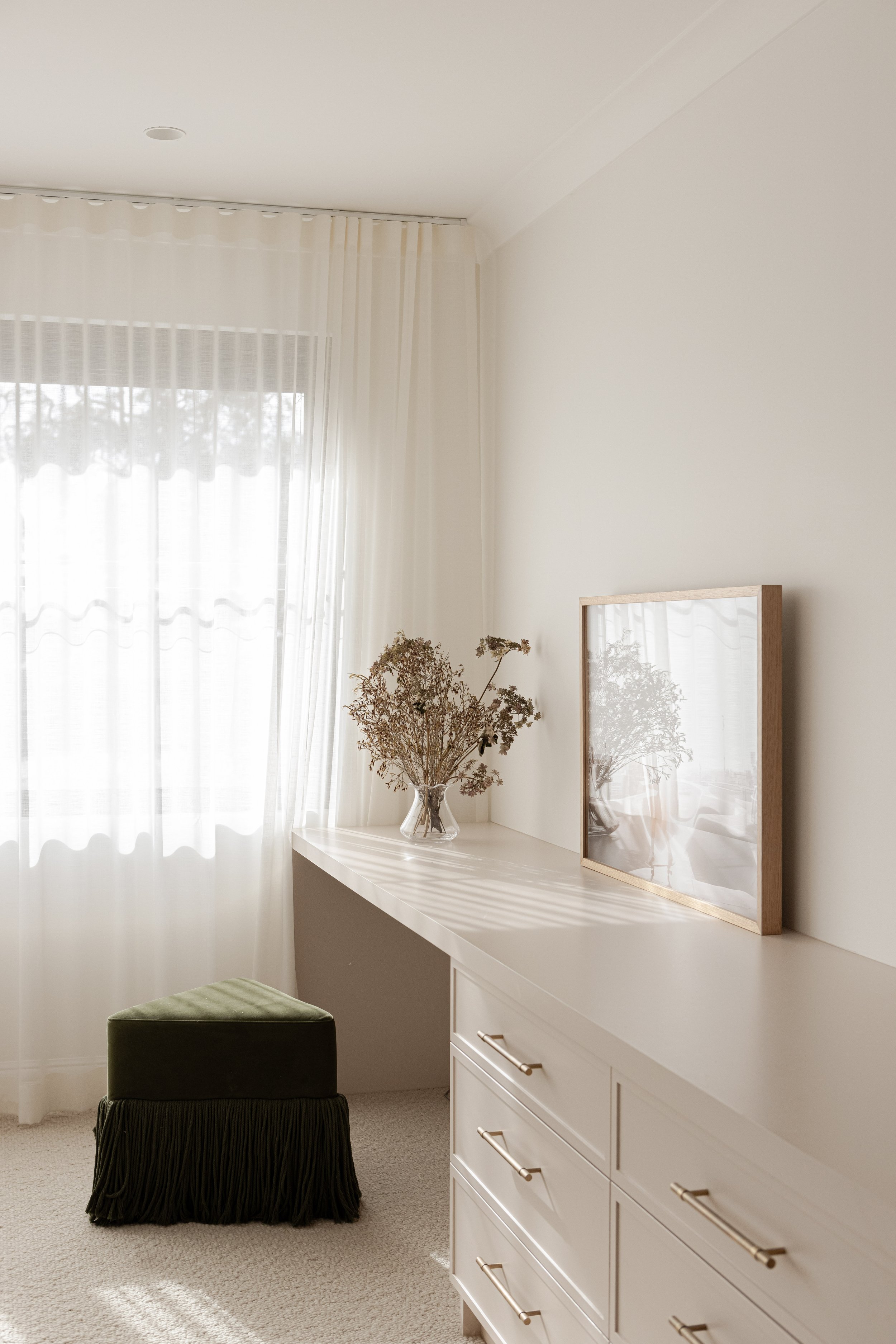 A bright, minimalist room corner with a white dresser, a framed mirror, a glass vase with dried flowers, and a green upholstered stool with a fringe skirt, near a window with sheer white curtains letting in natural light.