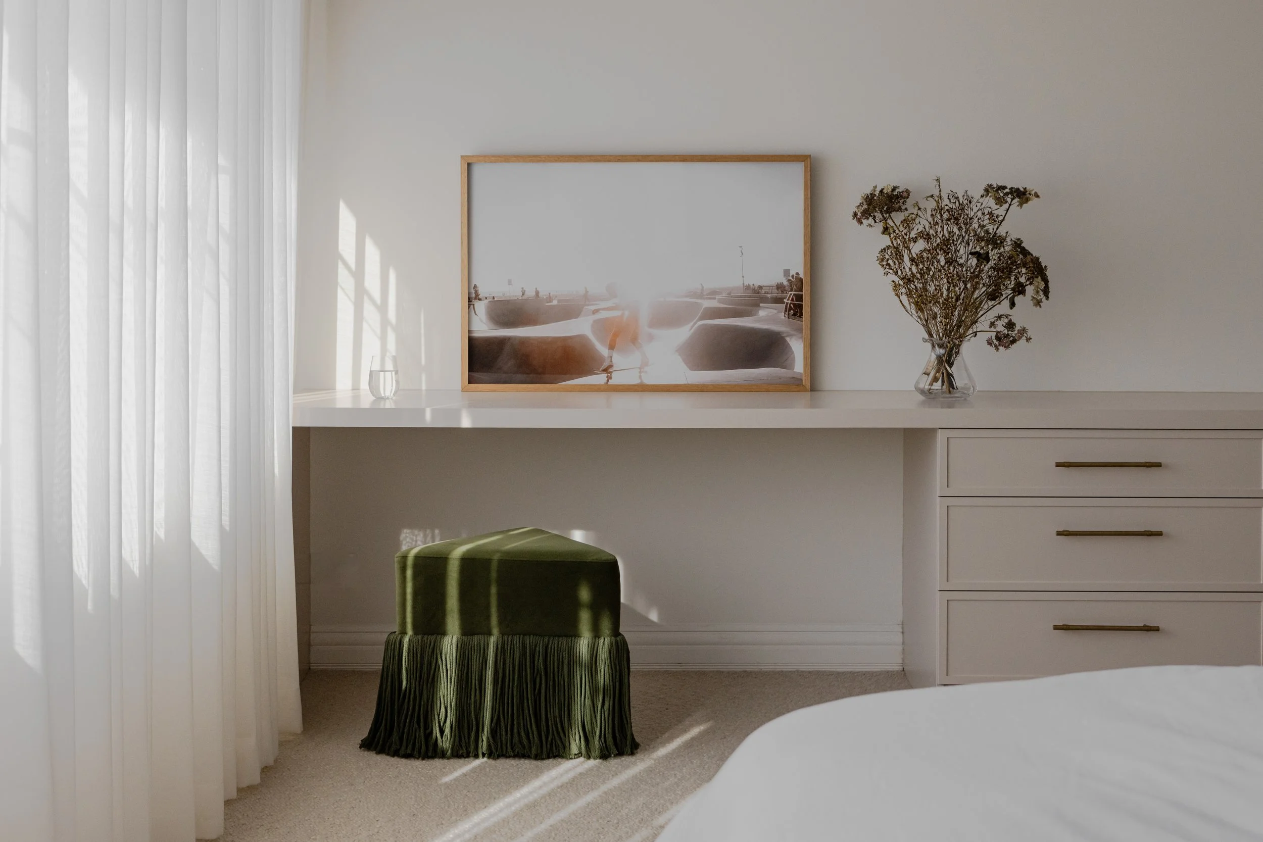 A bedroom corner with white curtains, a white desk against the wall, a framed artwork, a vase with dried flowers, a green ottoman with fringe, and a white drawer unit.