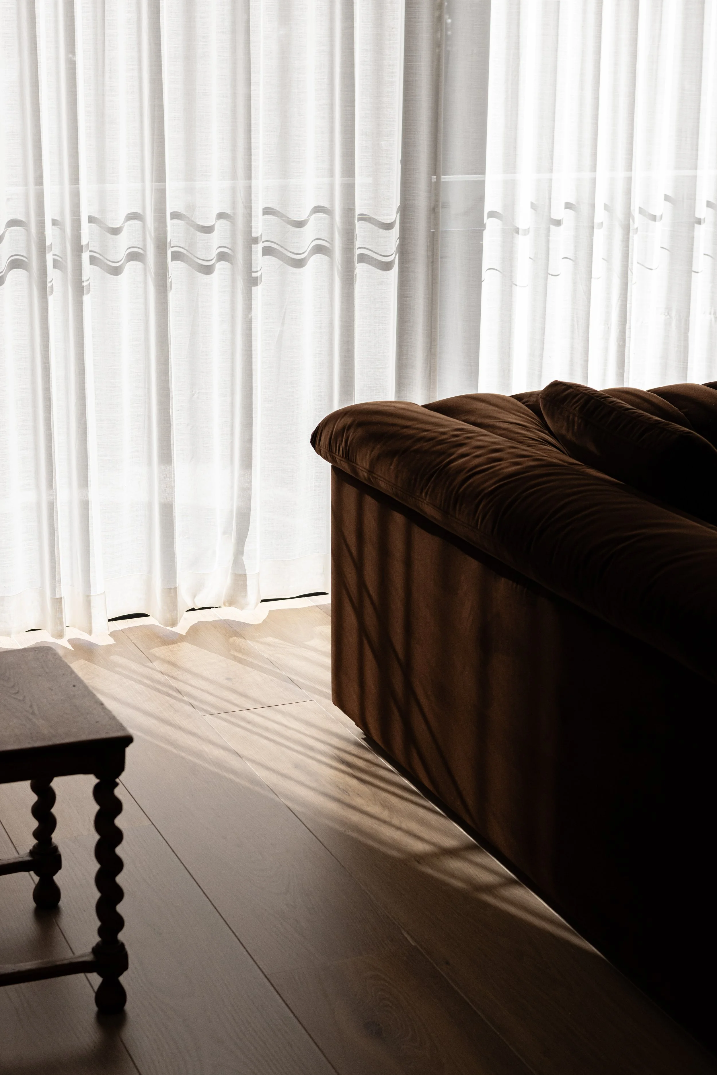 Sunlight filters through white curtains onto wooden floors and a brown couch in a living room.