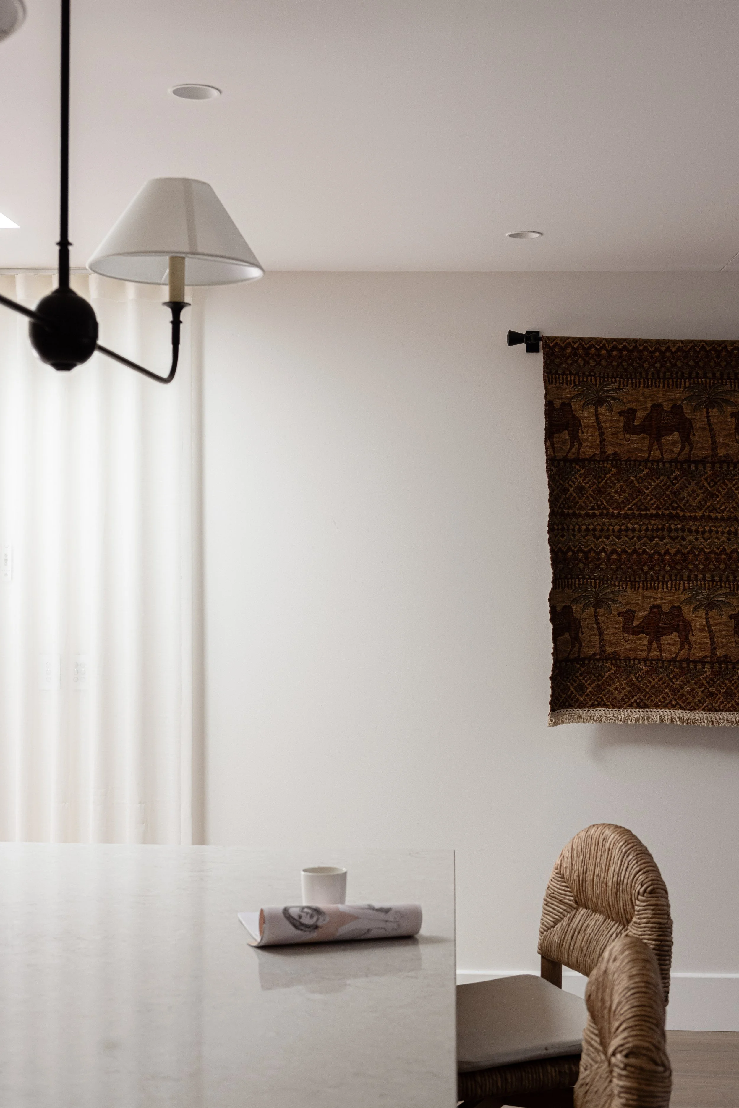 Interior dining room with a white wall, a tapestry with camels and palm trees, a woven chair, a white table with a cup and rolled paper, a black and white chandelier, and a window with white curtains.