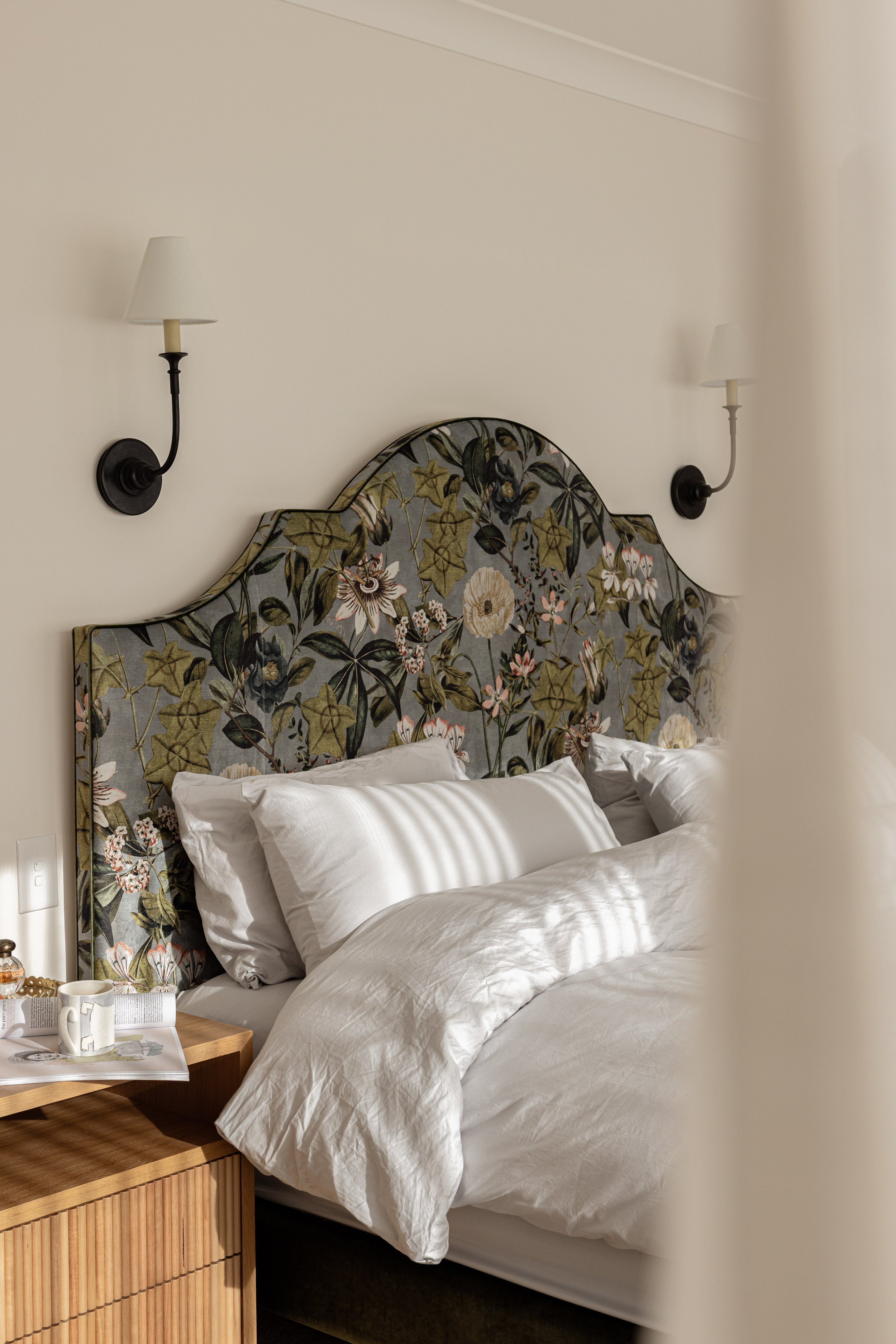 A neatly made bed with white pillows and bedding in a bedroom with wall-mounted lamps on a cream-colored wall behind a floral-patterned headboard.