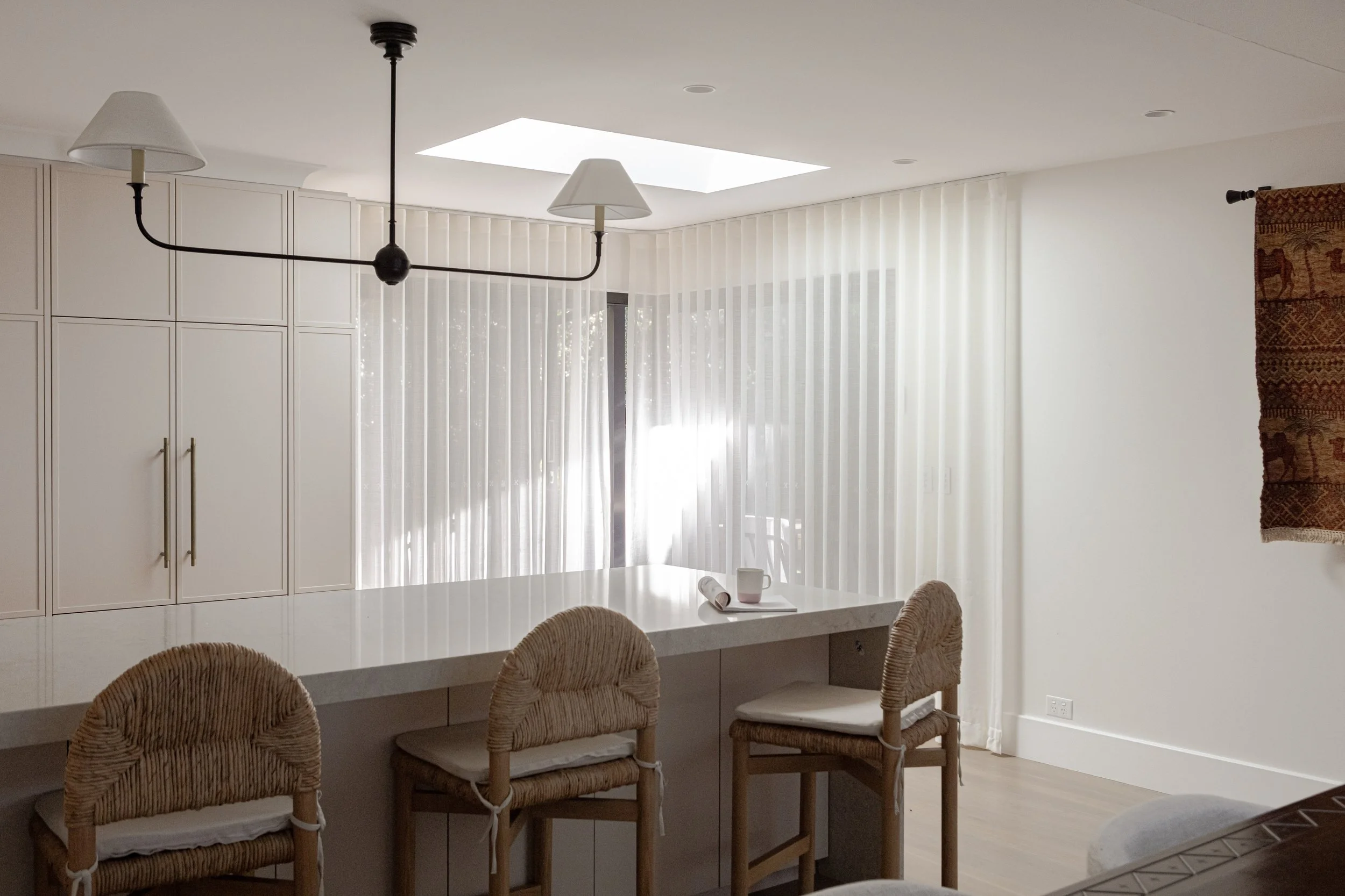 Modern kitchen with white cabinetry, a large island, wicker bar stools, vertical blinds, and a ceiling light fixture with two lampshades and a central black ball.