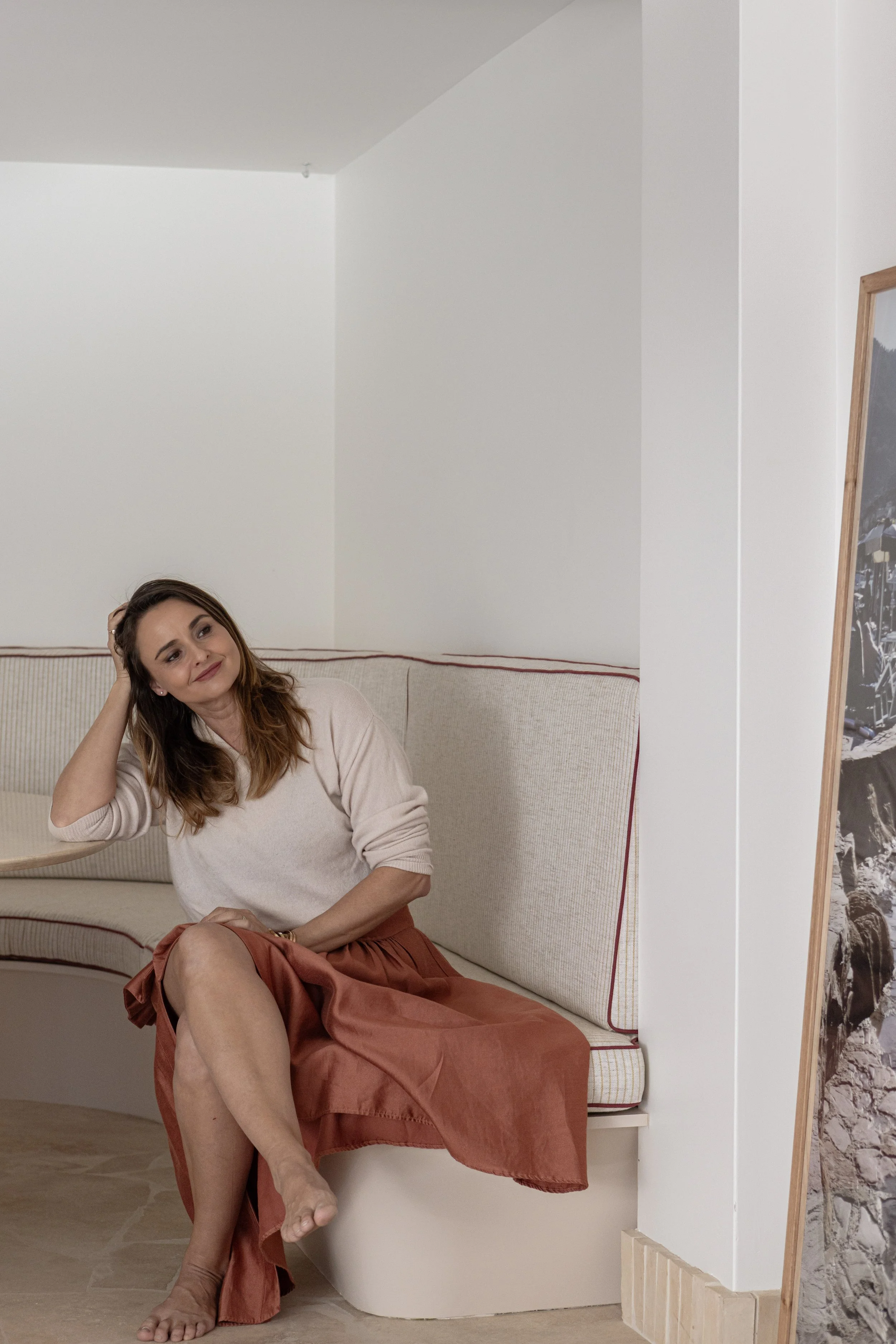 A woman with brown hair, wearing a beige sweater and a long orange skirt, sitting barefoot on a cream-colored curved sofa in a minimalistic room, with a painting or photograph partially visible on a stand to her right.