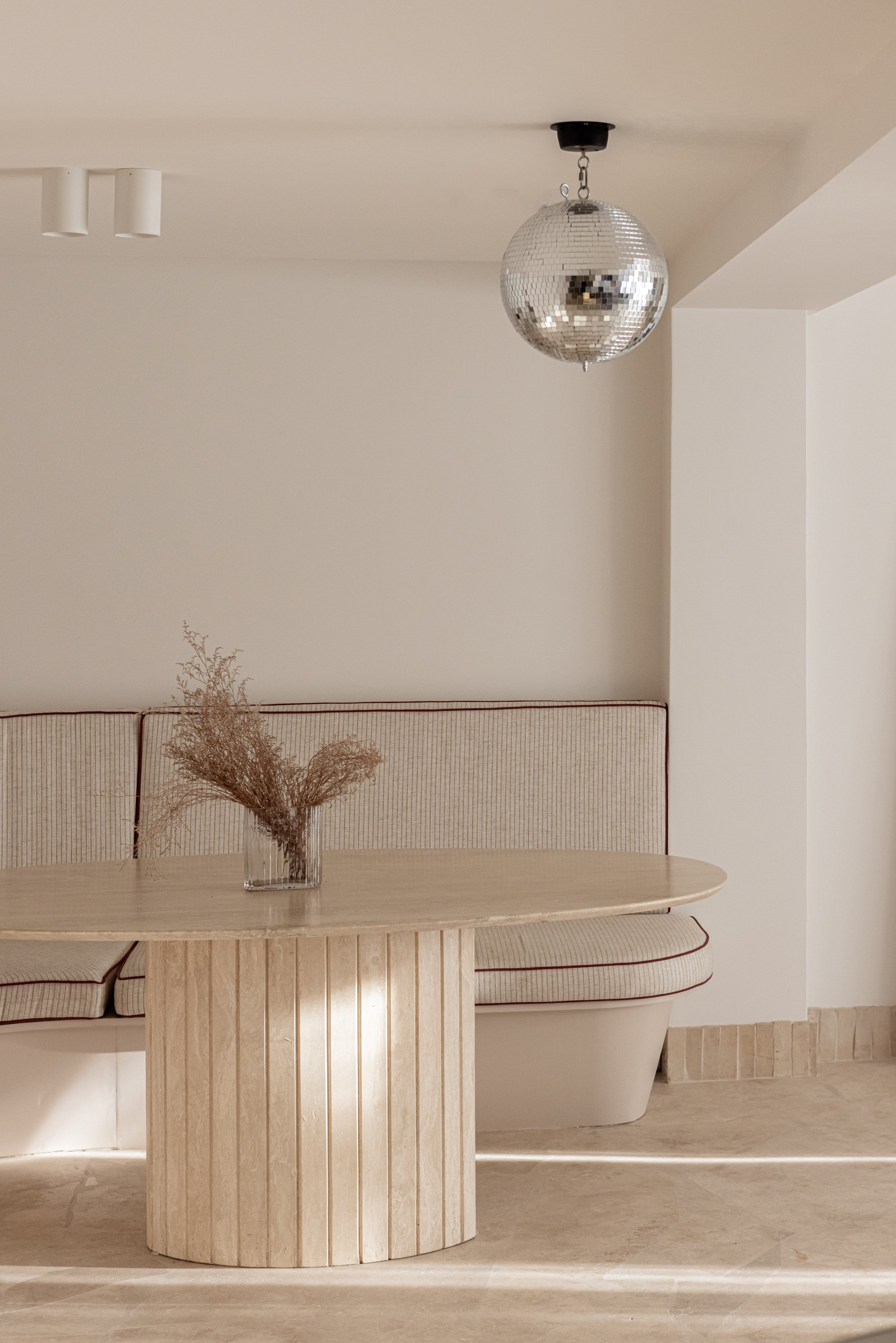 Minimalist dining area with a round wooden table, a built-in bench with beige striped upholstery, a glass vase with dried flowers, and a disco ball hanging from the ceiling.