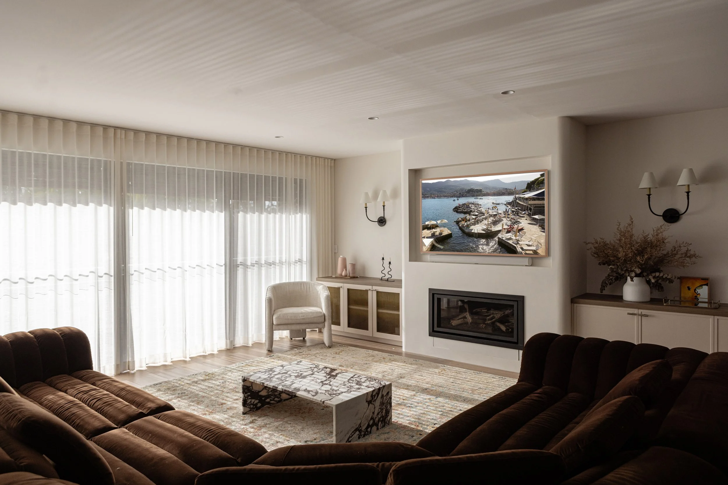 Living room with large windows, sheer curtains, a brown velvet sofa, a white armchair, a marble coffee table, a wall-mounted flat-screen TV showing a harbor scene, and a fireplace below the TV