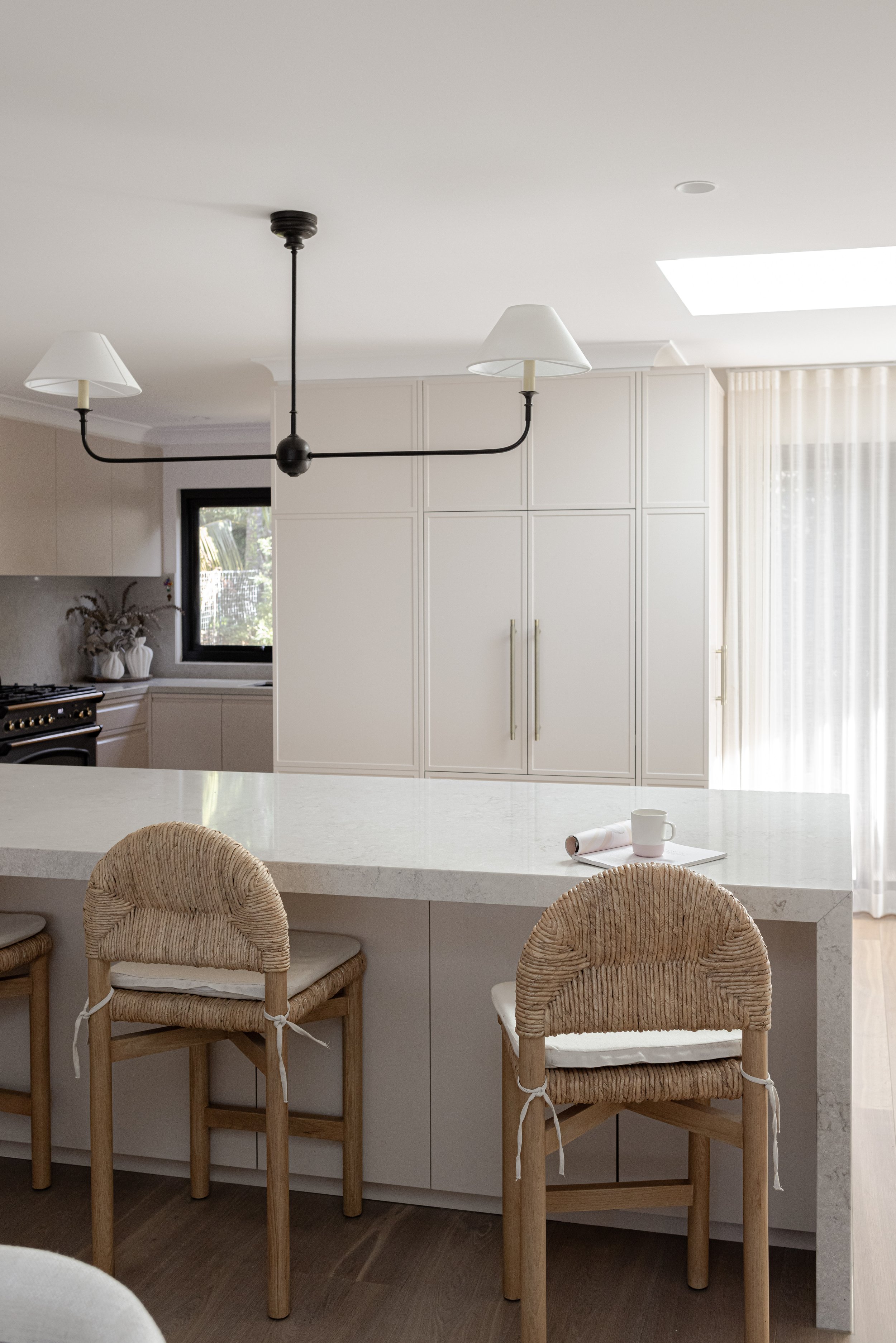 Modern kitchen with white cabinetry, a large white marble island, and natural light from a window and skylight. Bar stools with woven rattan seats and tied rattan backs are at the island. A black chandelier with white lampshades hangs from the ceilin