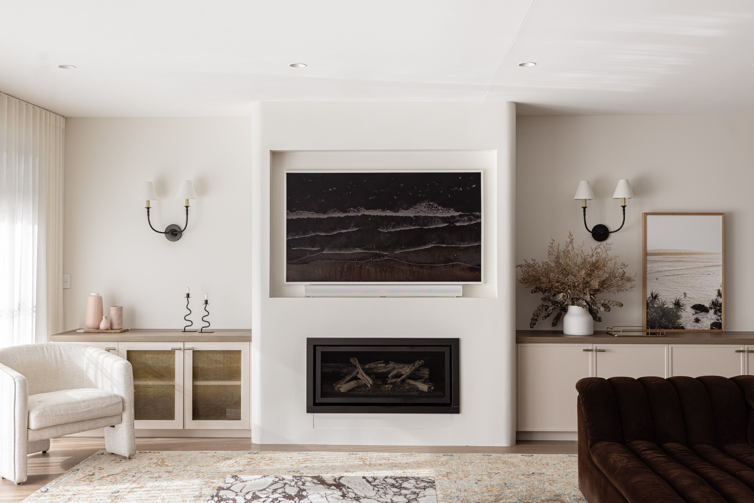 Modern living room with white walls, wall sconces, a large flat-screen TV above a fireplace, and decorative vases and artwork