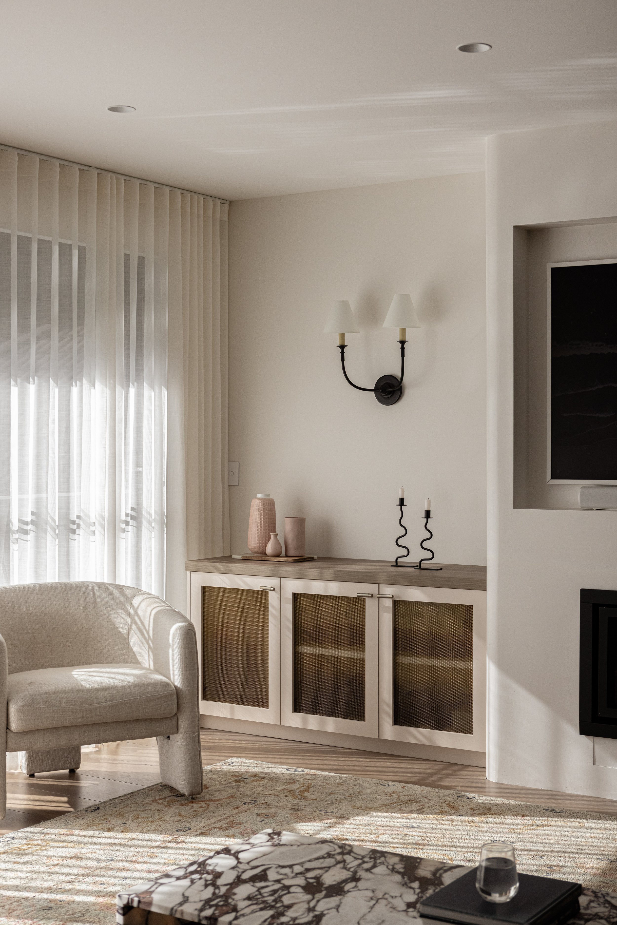 Living room with a beige armchair, a white cabinet with glass doors, a marble-topped coffee table, and a window with sheer curtains. Decorative items include vases and candle holders on the cabinet, and wall-mounted lamps.