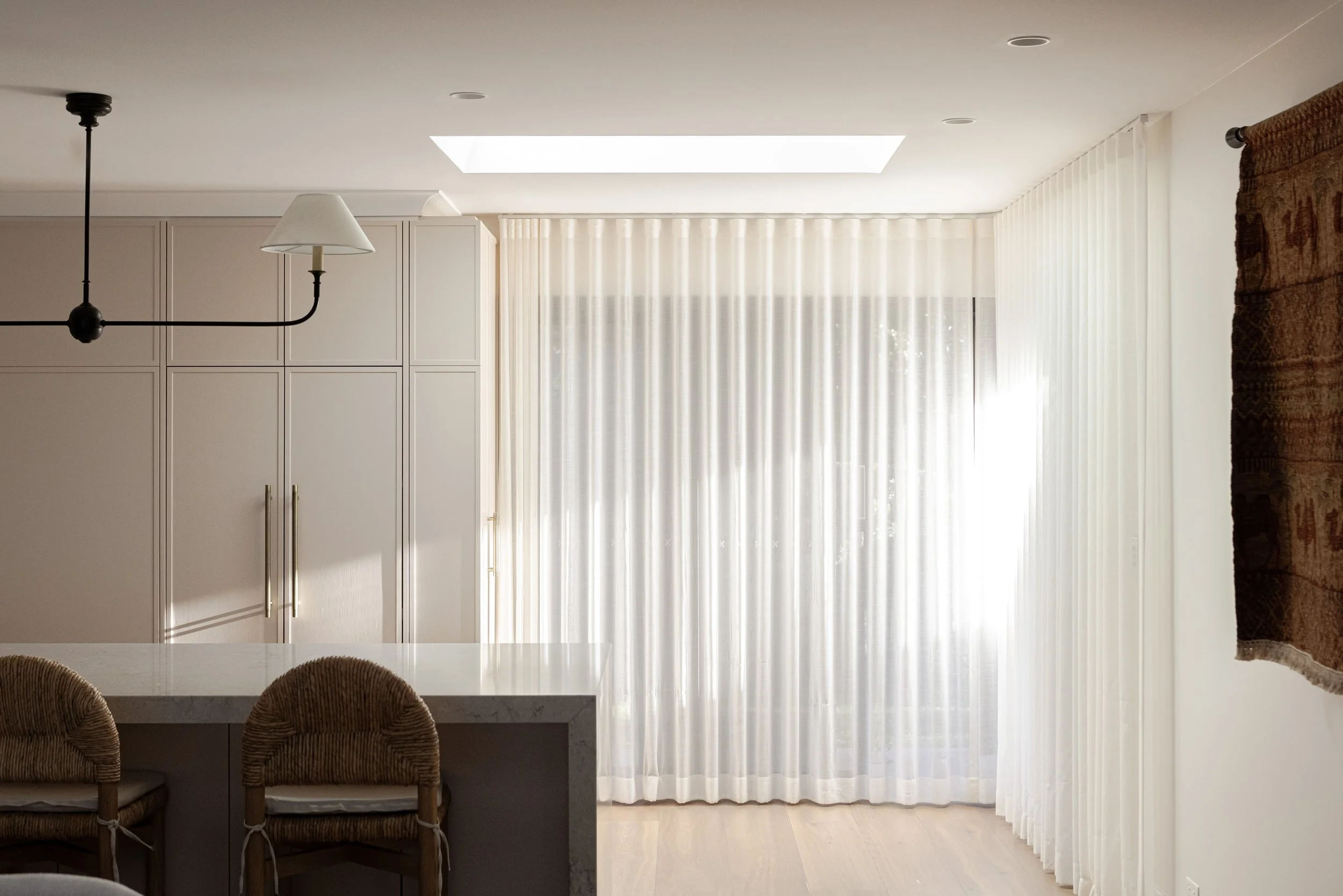 Bright kitchen with white cabinets, large window, sheer curtains, and a marble island with two rattan chairs.
