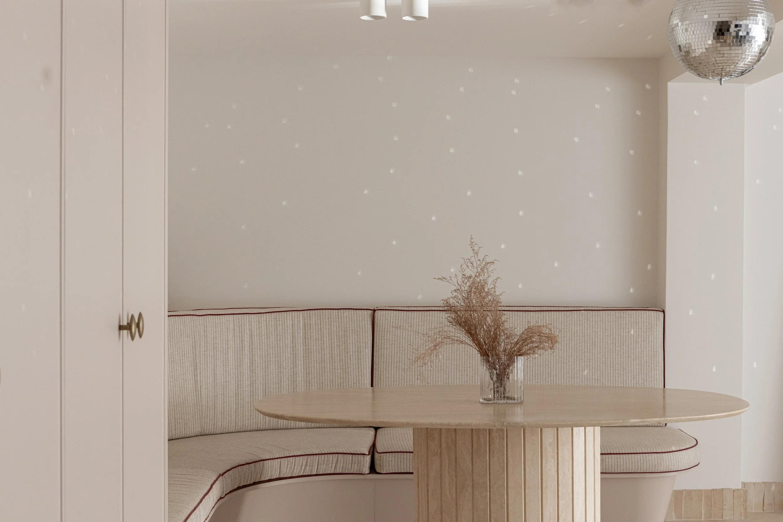 Minimalist dining nook with a curved beige upholstered banquette, a round wooden table, a glass vase with dried flowers, and a disco ball hanging from the ceiling, set against a plain white wall with small light reflections.
