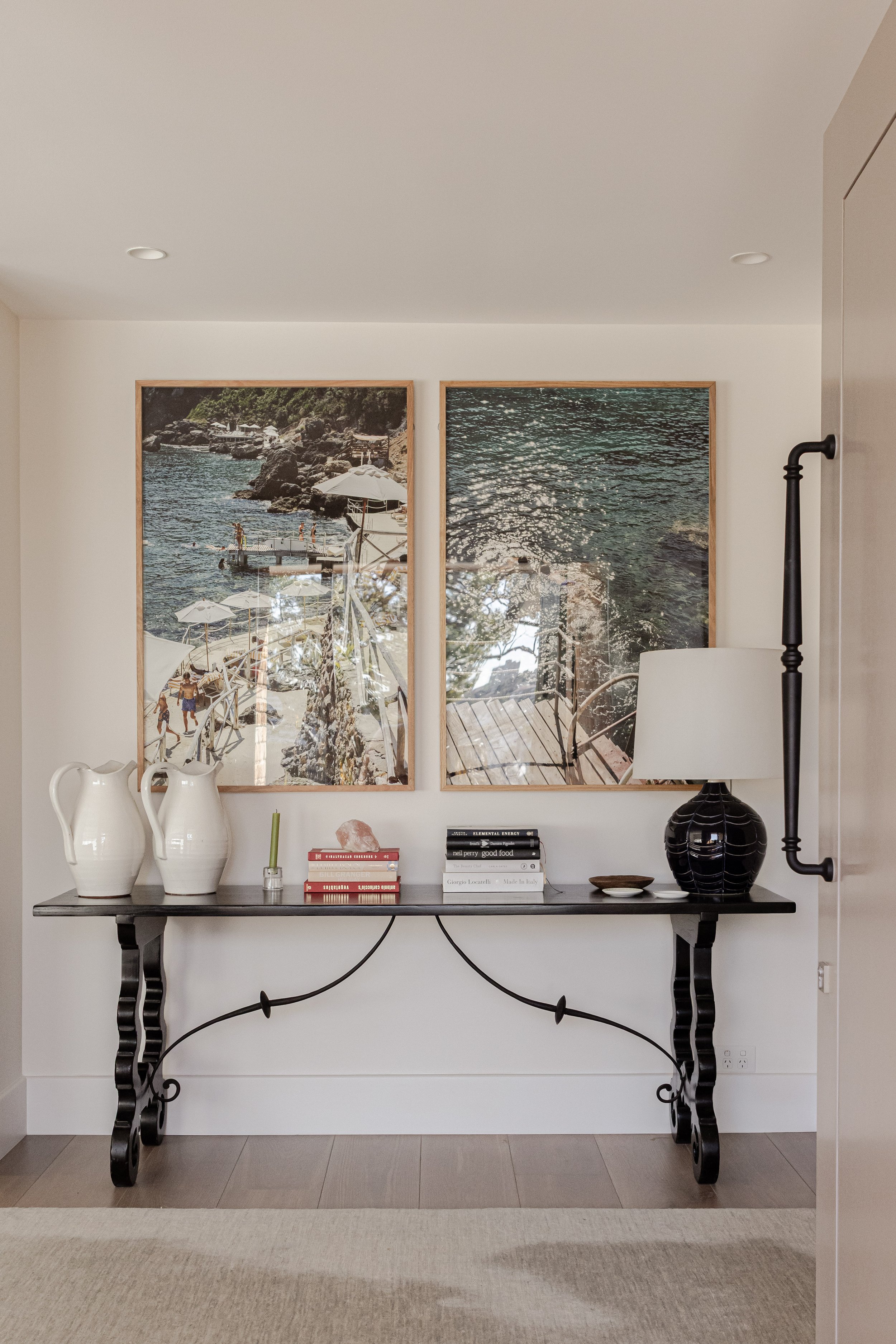 A black console table with decorative iron legs, positioned against a white wall. On the table are two white pitchers, a small green plant, a pink salt lamp, and several stacked books. To the right of the table is a black and white ceramic table lamp