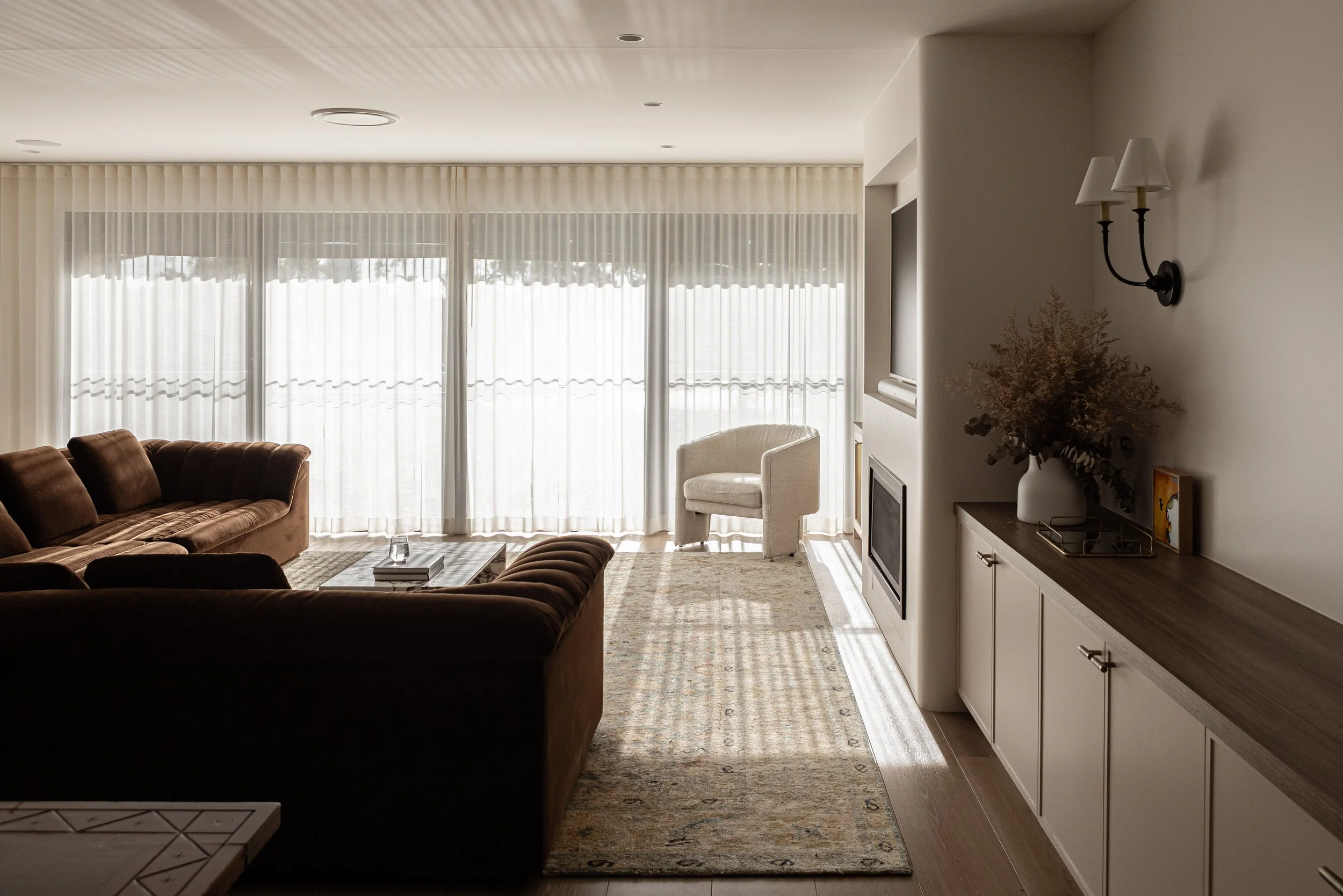 A spacious living room with large windows covered by sheer curtains, brown sofas, a coffee table, a white armchair near the window, a built-in fireplace, a wooden sideboard with a white vase of dried flowers, and wall-mounted light fixtures.