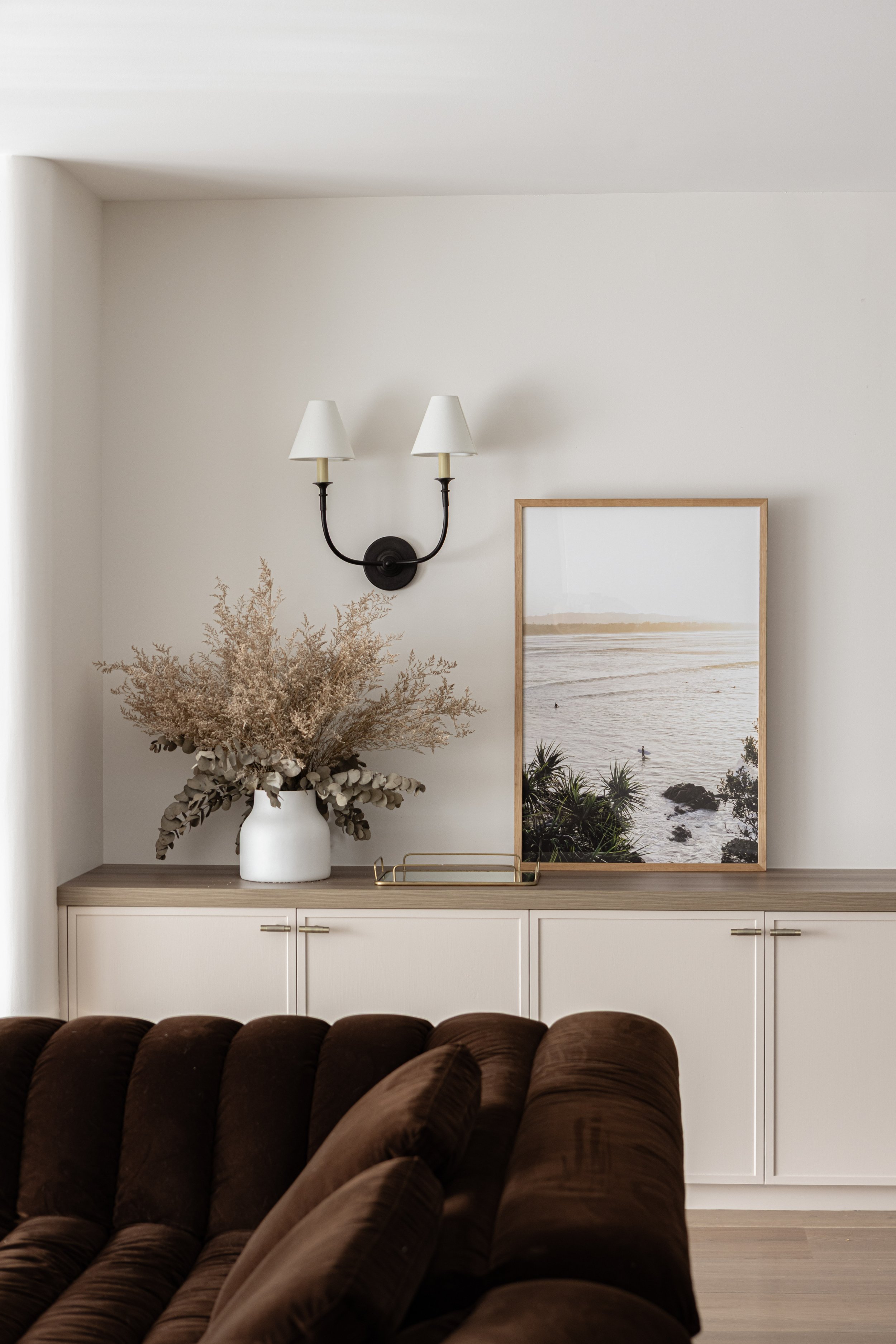Living room corner with a sideboard, a large white vase with dried flowers, a framed beach photograph, and a wall-mounted double-arm lamp with white shades.