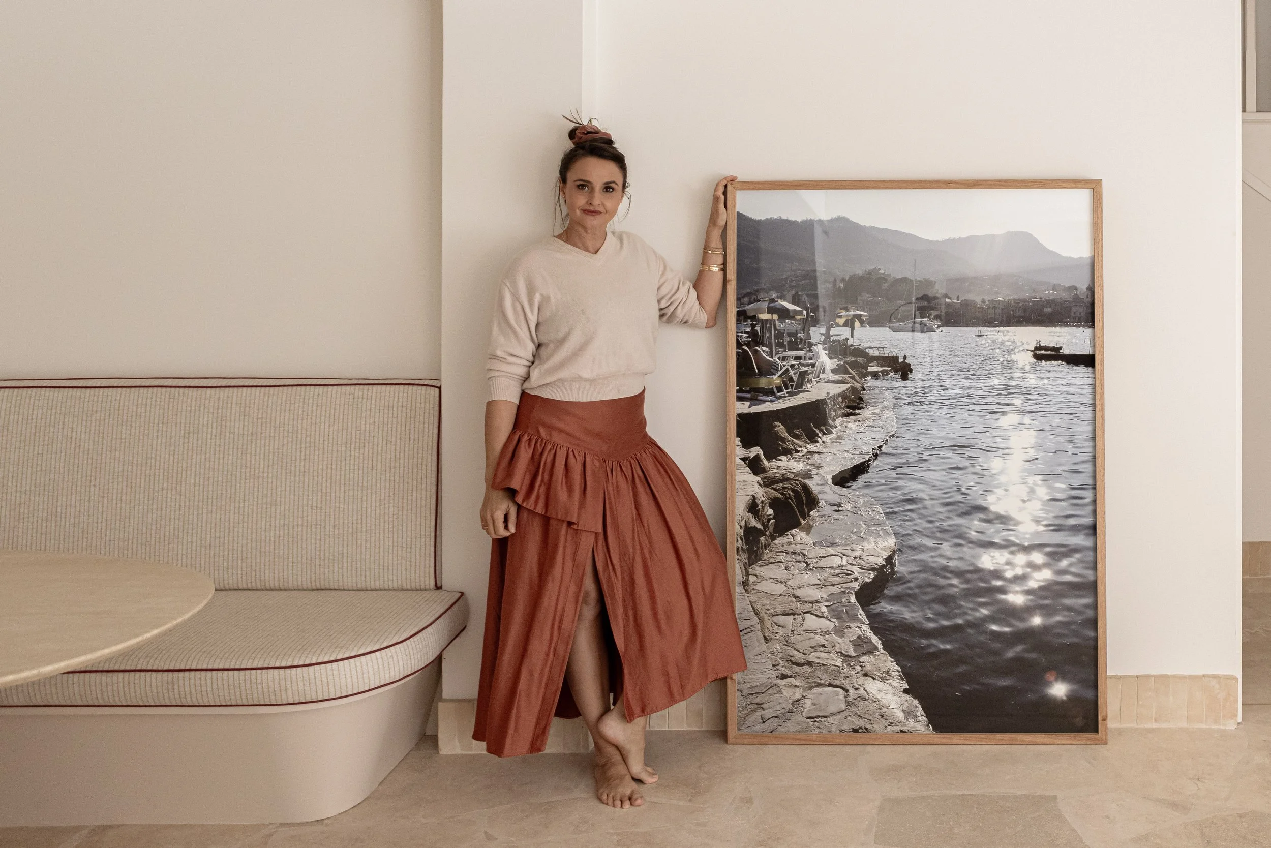 Woman with brown hair in a bun, wearing a beige sweater and rust-colored skirt, standing barefoot next to a framed photo of a lakeside scene with mountains, water, and boats.
