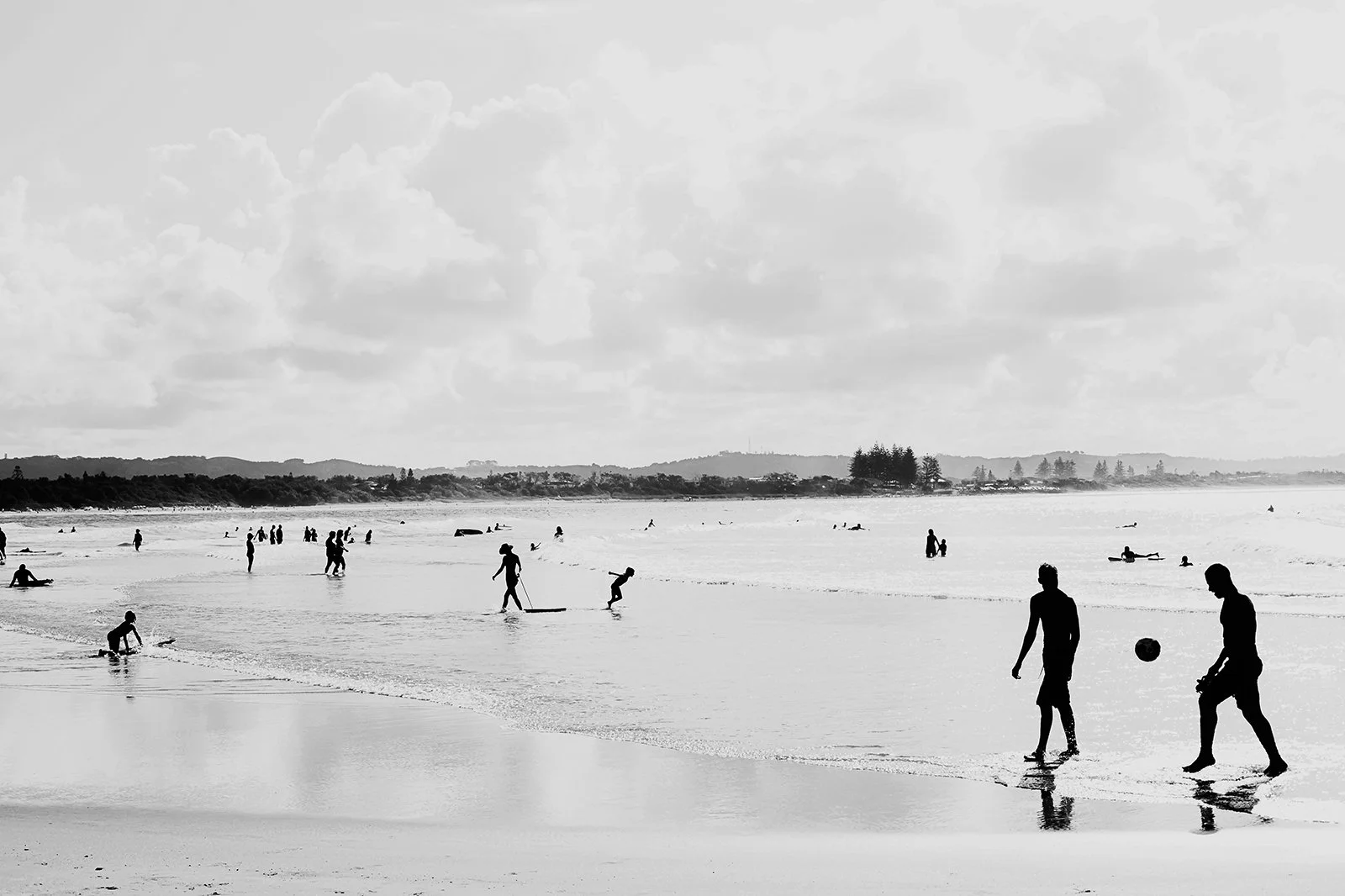 People enjoying a day at the beach, some walking along the shoreline, others surfing or playing in the water, clouds in the sky, distant land across the water.