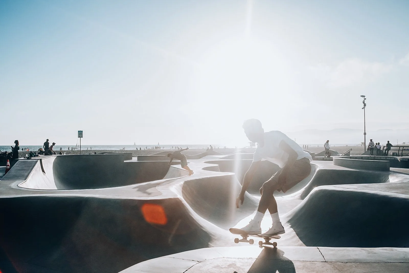 A skateboarder performing a trick at a skate park near the beach with people walking and relaxing in the background, under bright sunlight.