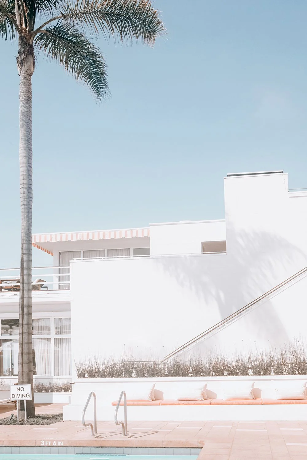 A white building with modern architecture, a large palm tree, a clear blue sky, and an outdoor lounge area with pink cushions near a swimming pool.