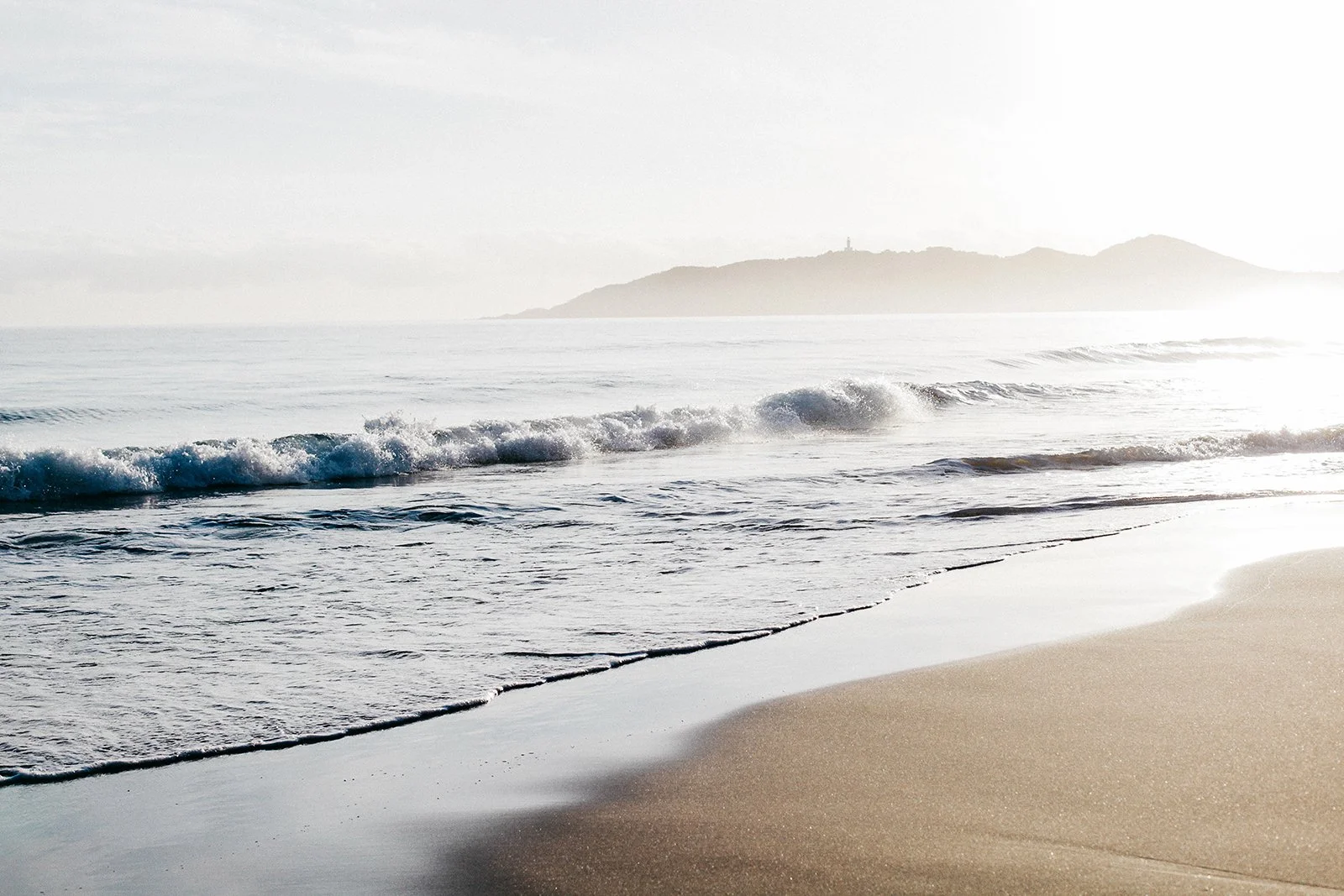 A calm beach with gentle waves and a distant hilly island in the background.