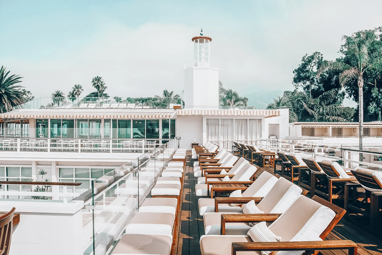 Empty outdoor rooftop lounge with lounge chairs, pillows, and a lighthouse structure, with palm trees and blue sky in the background.