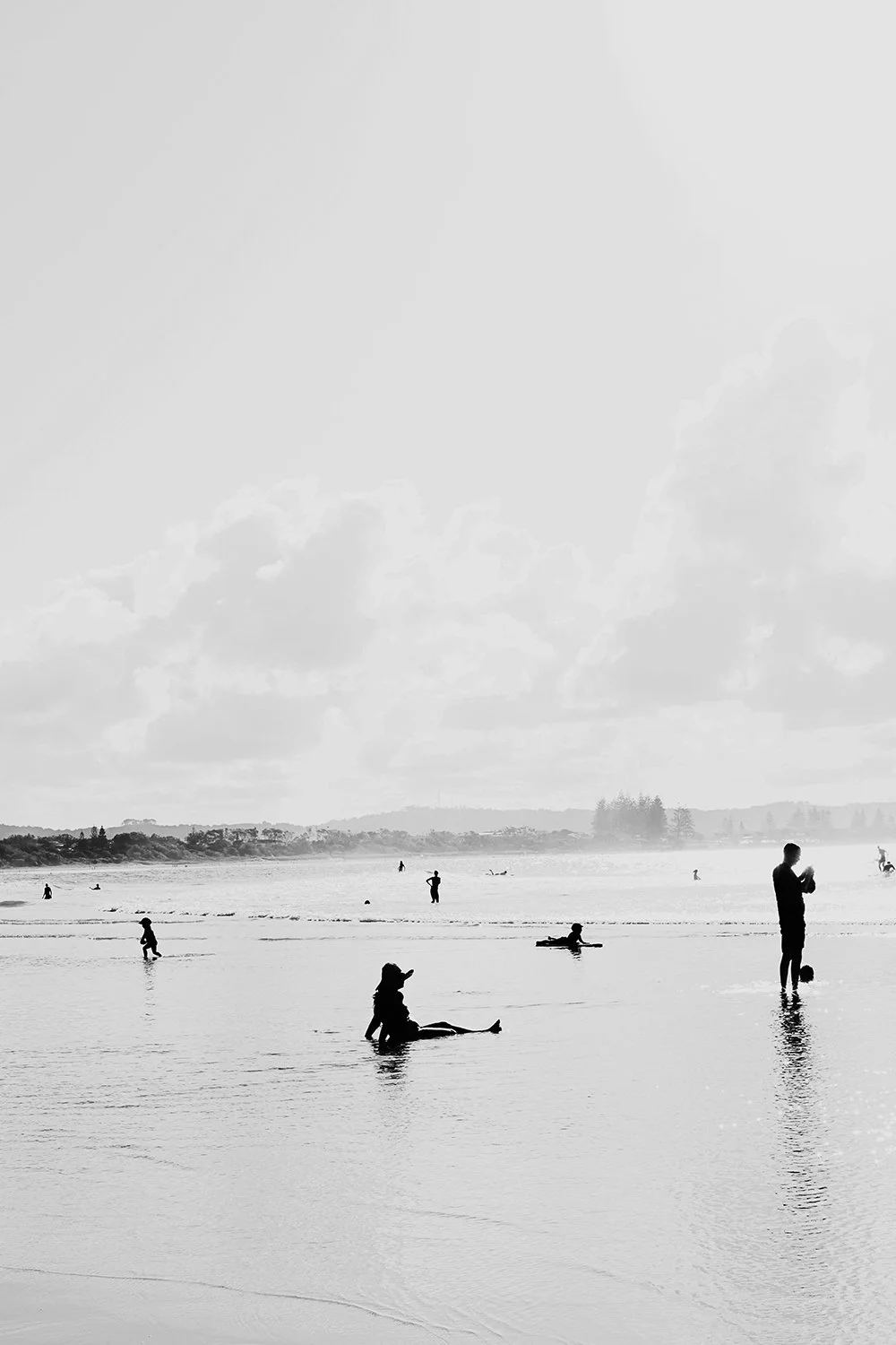 People at the beach, some wading in shallow water, some standing or sitting, in a black-and-white photo with a cloudy sky.