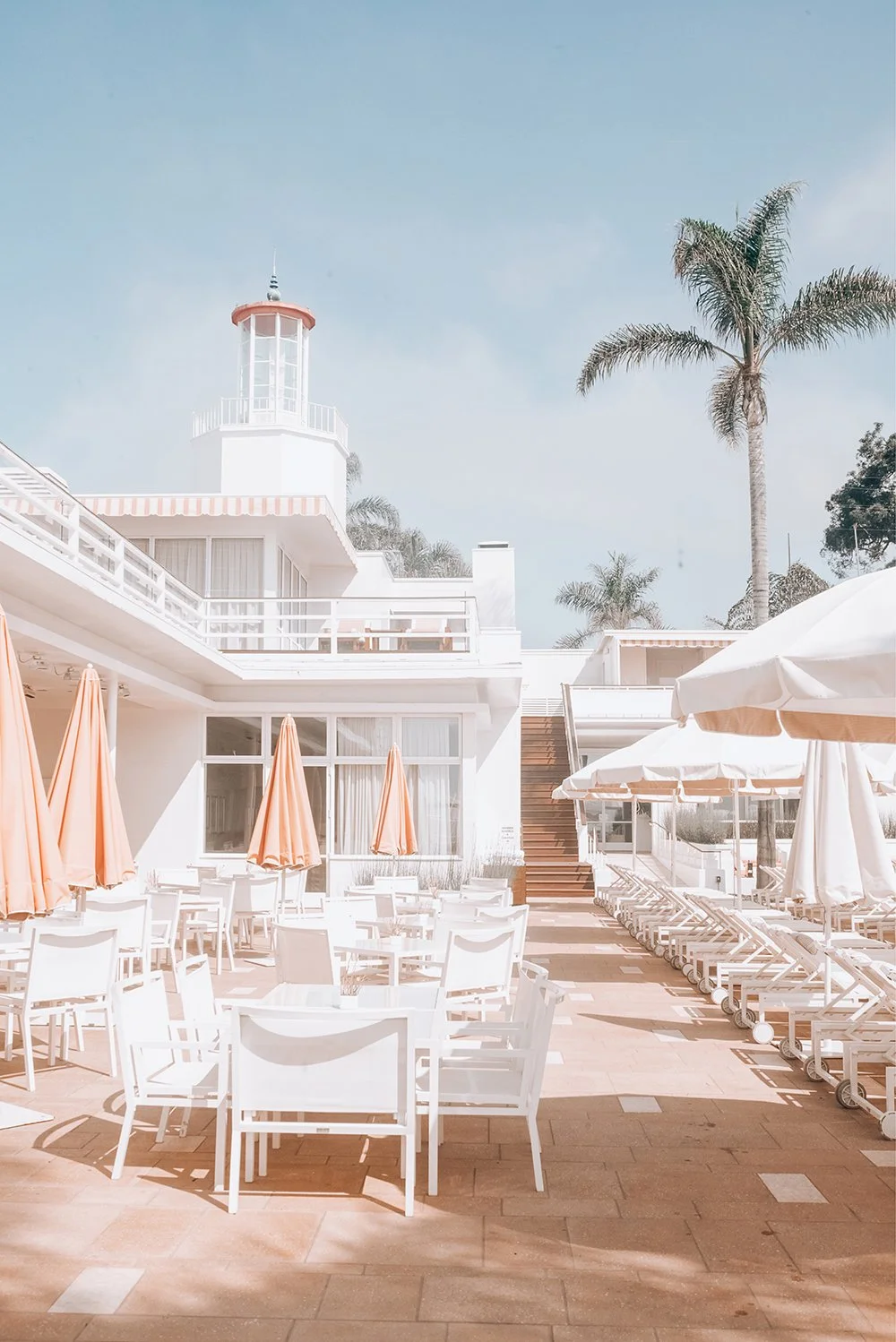 Outdoor poolside area with white chairs, umbrellas, and a white building with a lighthouse and palm trees in the background