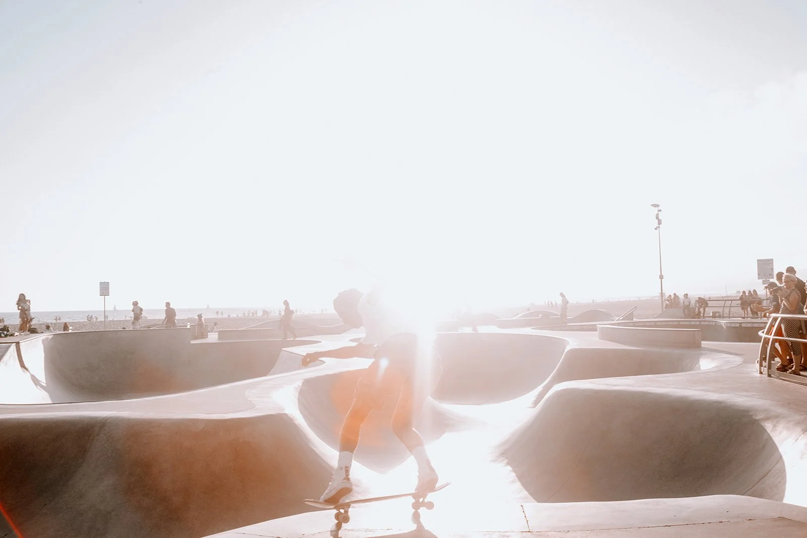 Skateboarder performing tricks at a skate park near the beach with people walking and enjoying the sunny day.