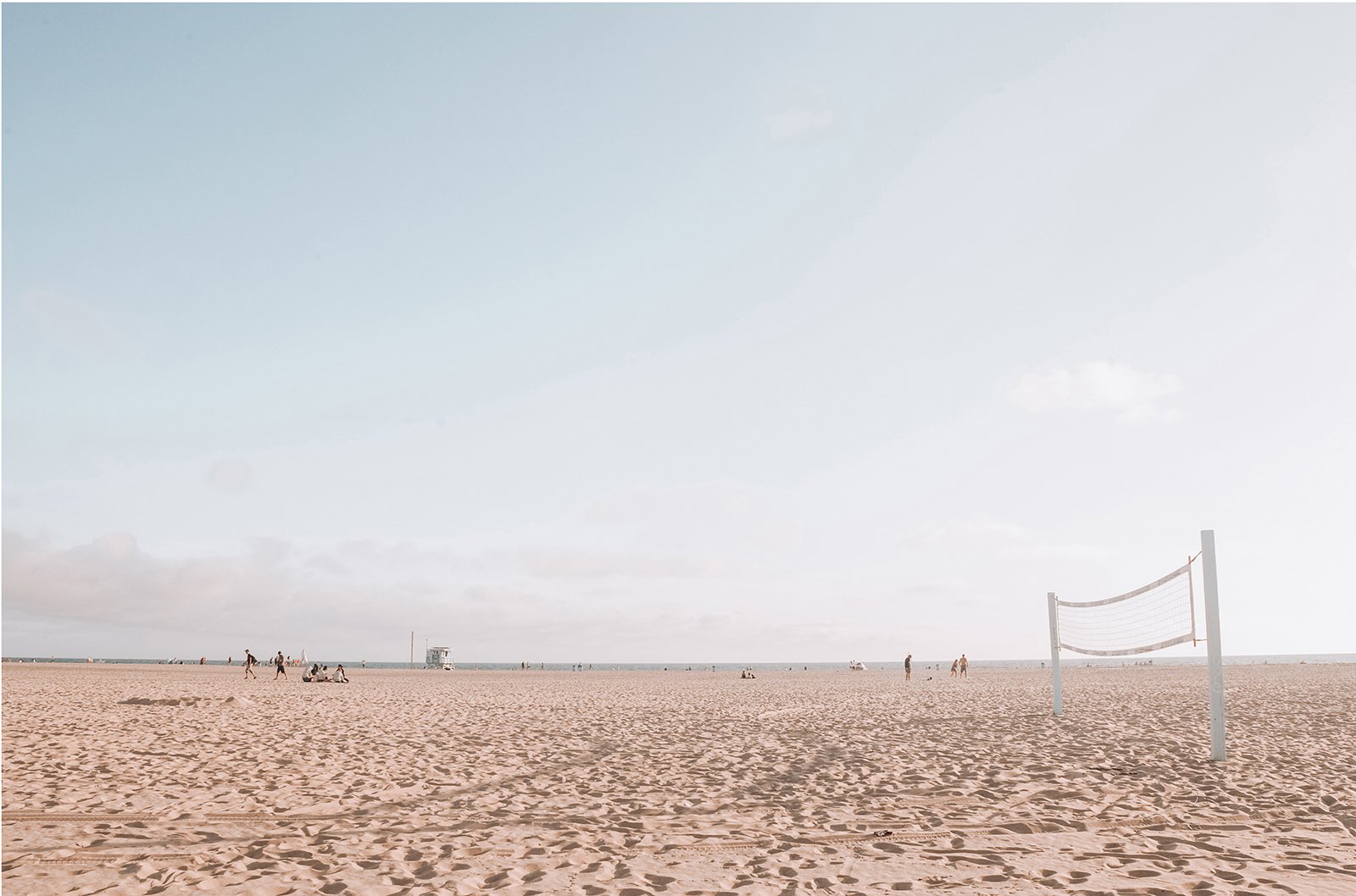 A sandy beach with a volleyball net on the right, a few people sitting and walking, and a lifeguard tower in the distance, under a partly cloudy sky.