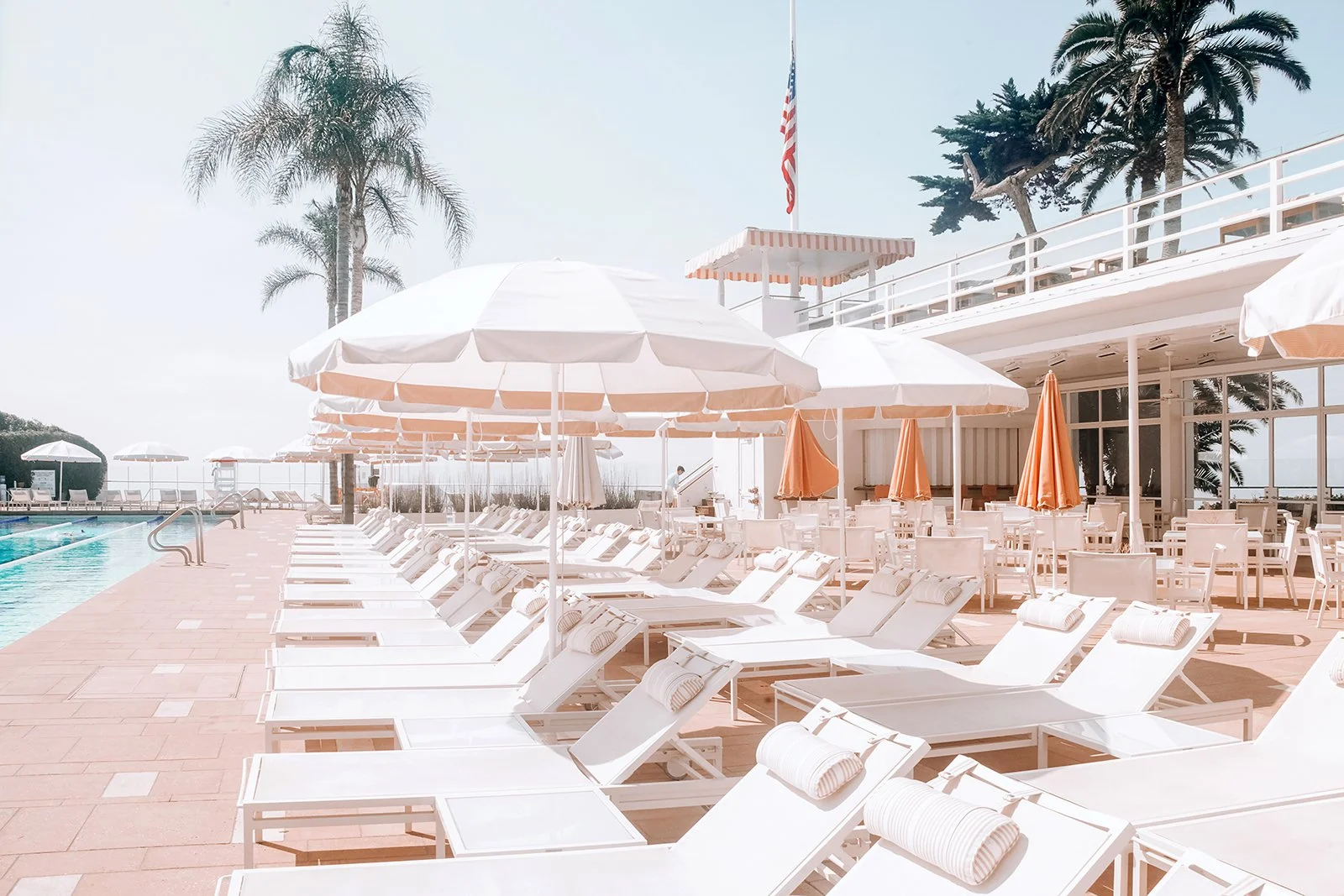 Empty poolside area with white lounge chairs, umbrellas, and towels, overlooking a pool at a sunny resort with palm trees.
