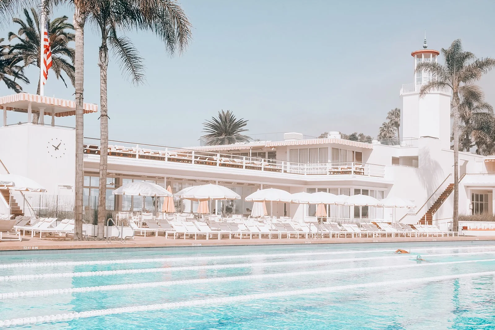 A swimming pool area with white lounge chairs and umbrellas, surrounded by tall palm trees, and a white building with a tower in the background.