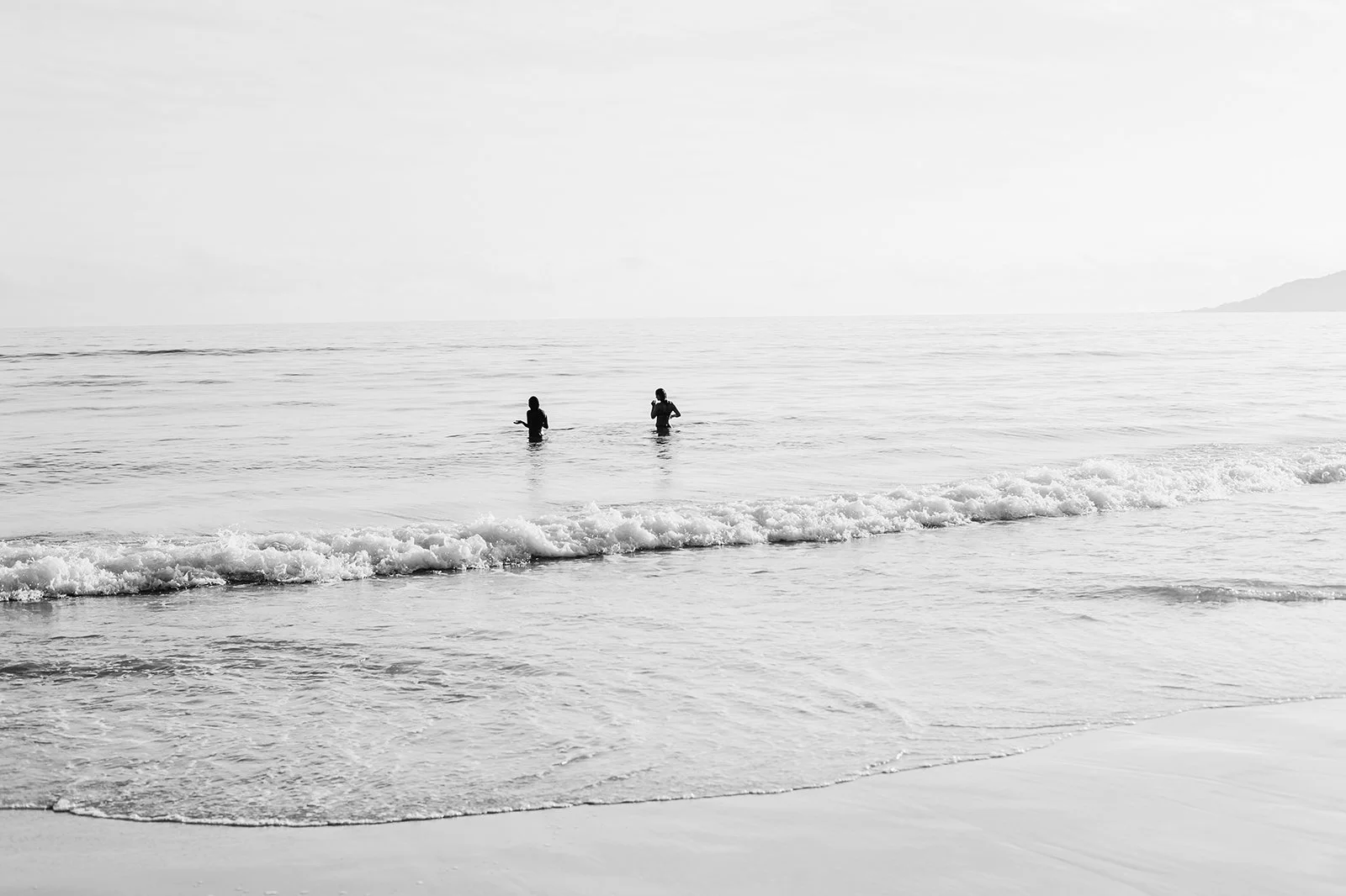 Two people standing in the ocean water near the shoreline, with gentle waves and a distant foggy horizon.
