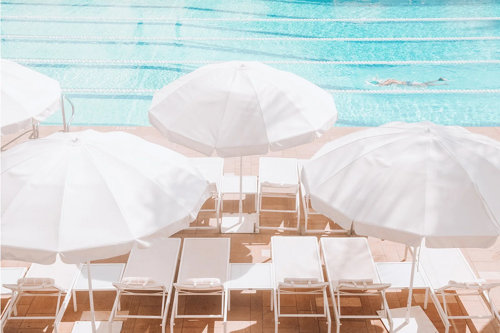 Empty poolside lounge chairs with white umbrellas, pool with swimmers in the background