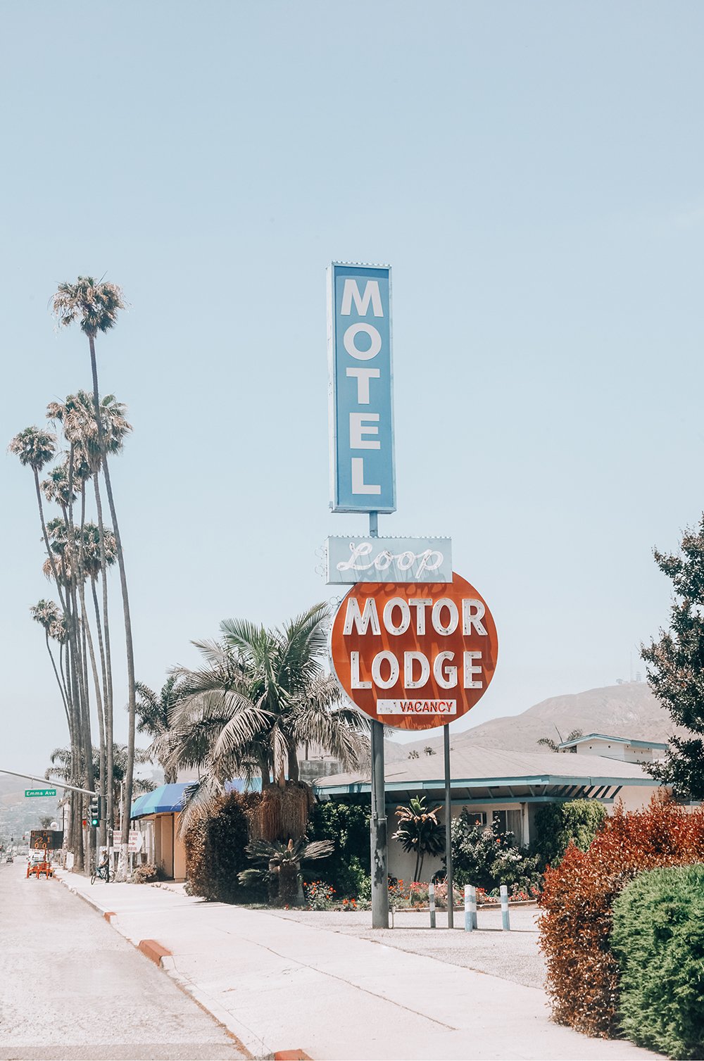 A street view with tall palm trees, a motel sign reading 'Loop Motor Lodge', and a building surrounded by bushes and trees under a clear sky.