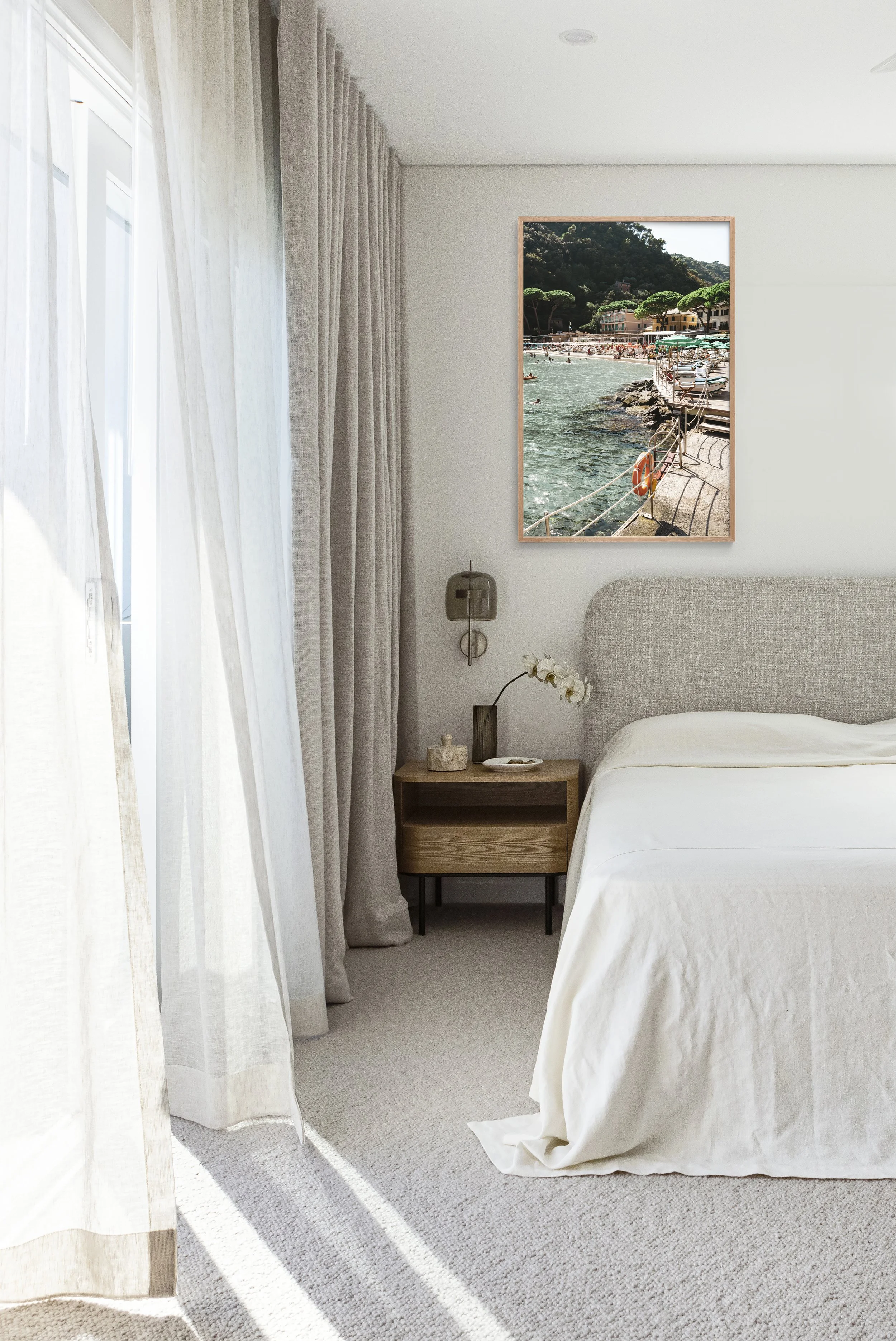 A bedroom with a nightstand, a bed with a beige headboard, and a framed photo of a seaside scene on the wall. Light-colored curtains are blown by the breeze near a window.