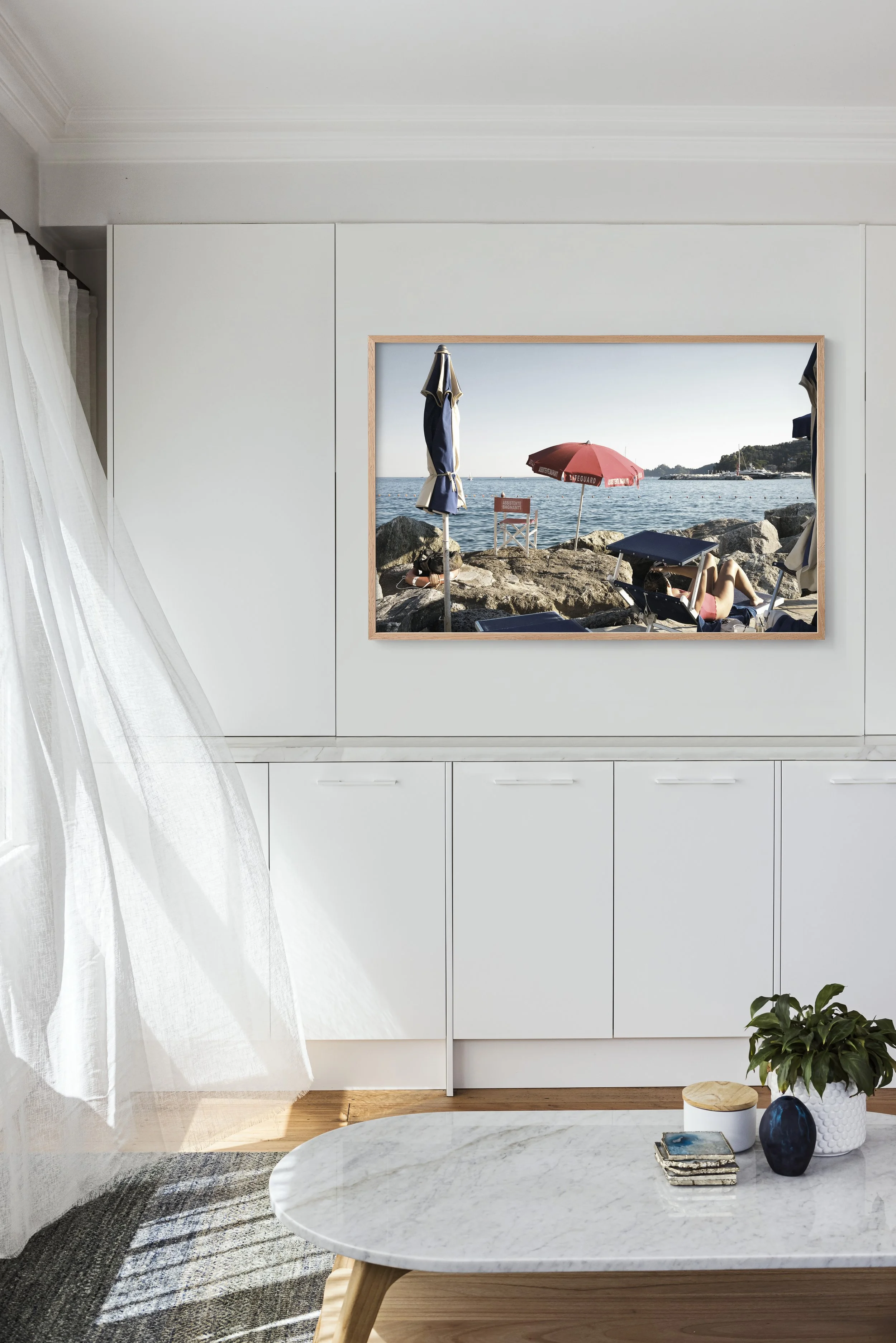 Living room interior with a framed seaside landscape painting on a white wall, sheer curtains on a window on the left, a marble-topped coffee table with decorative objects, and a potted plant on a wooden floor.