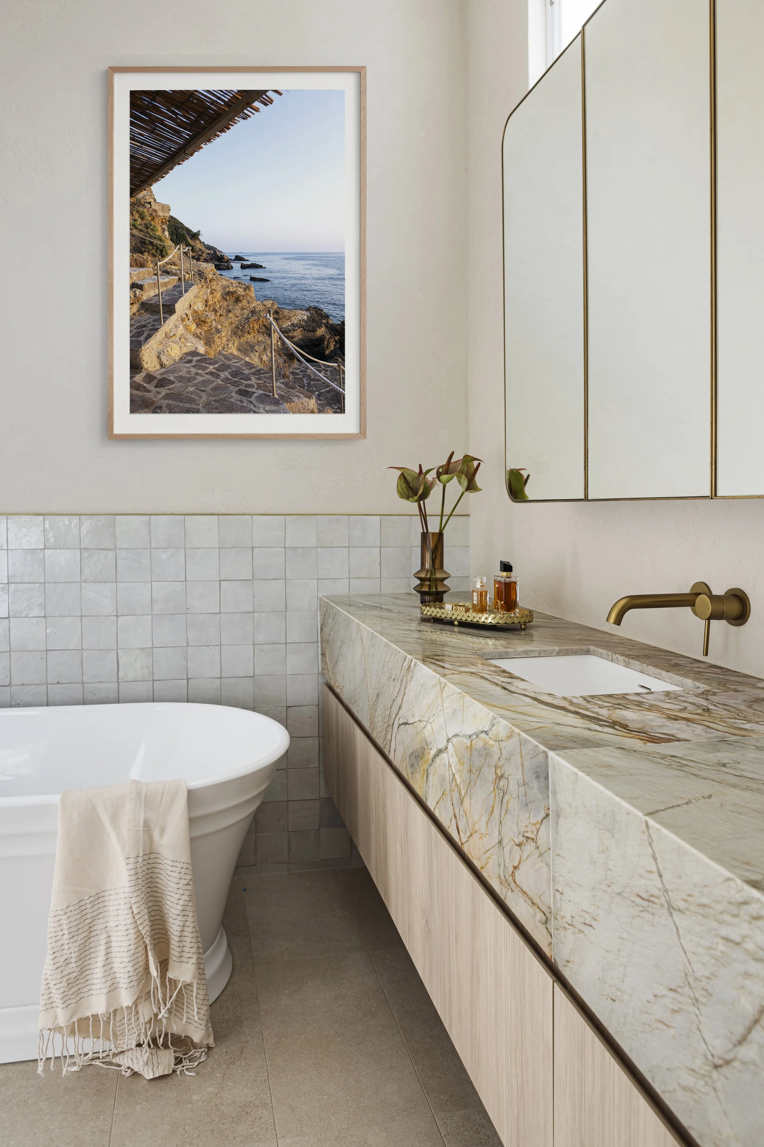 Interior bathroom with a marble countertop, a vase with lotus flowers, skincare bottles, a large framed ocean view photograph, and a white bathtub with a towel hanging over its edge.