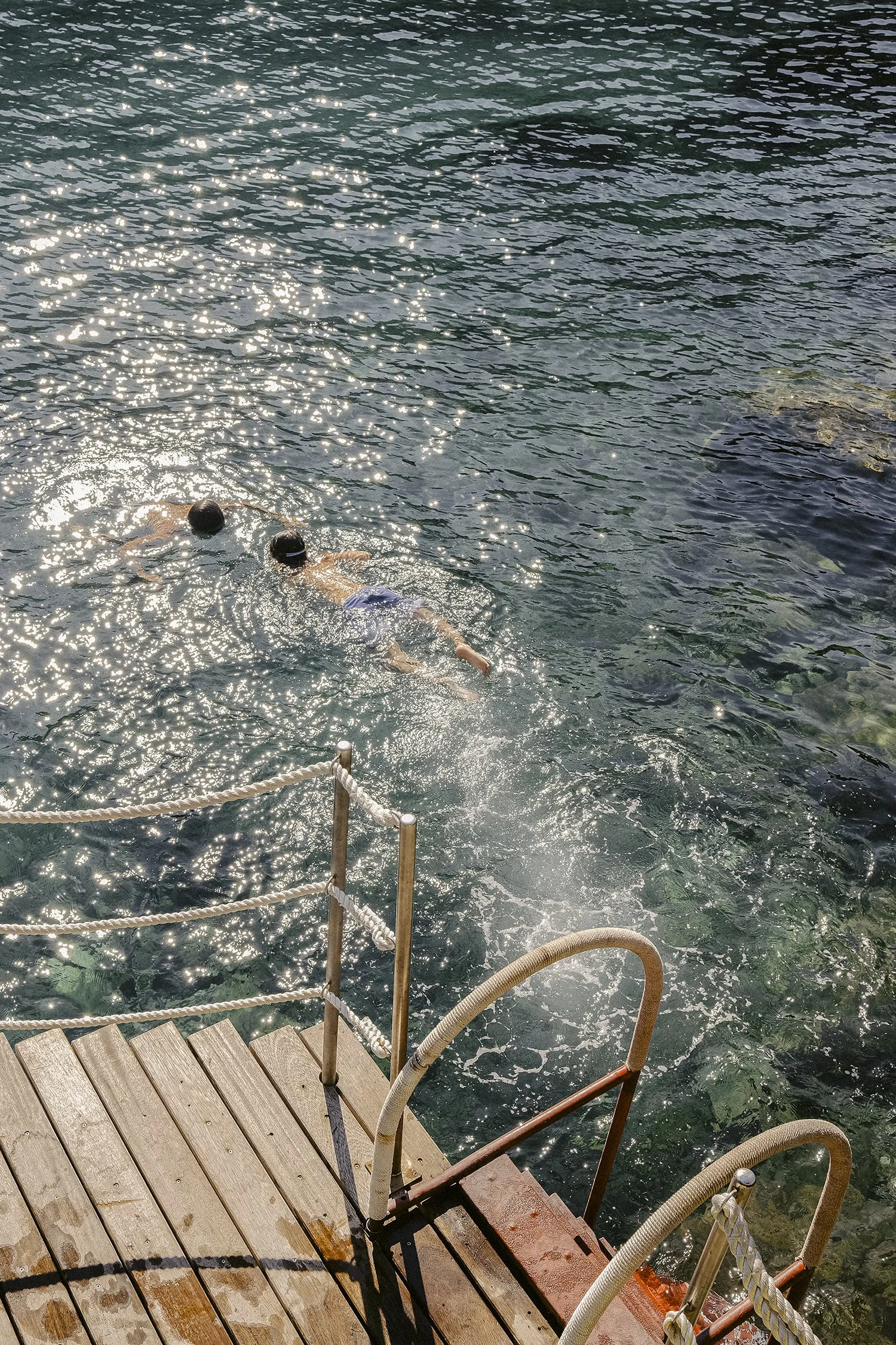 Two children swimming in a body of water near a wooden dock with ropes, sunlight reflecting on the water, and rocks visible beneath the surface.