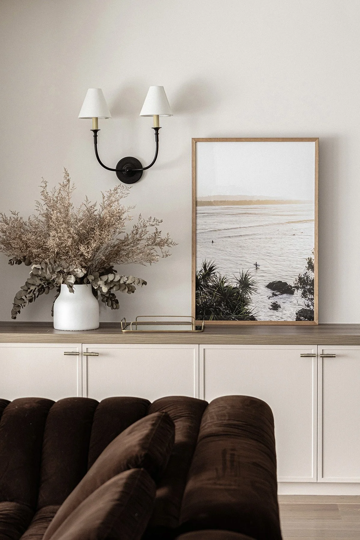 Interior of a living room with a brown couch, a wooden sideboard, a framed photo of a beach scene, a white vase with dried flowers, and a wall-mounted light fixture with two lampshades.