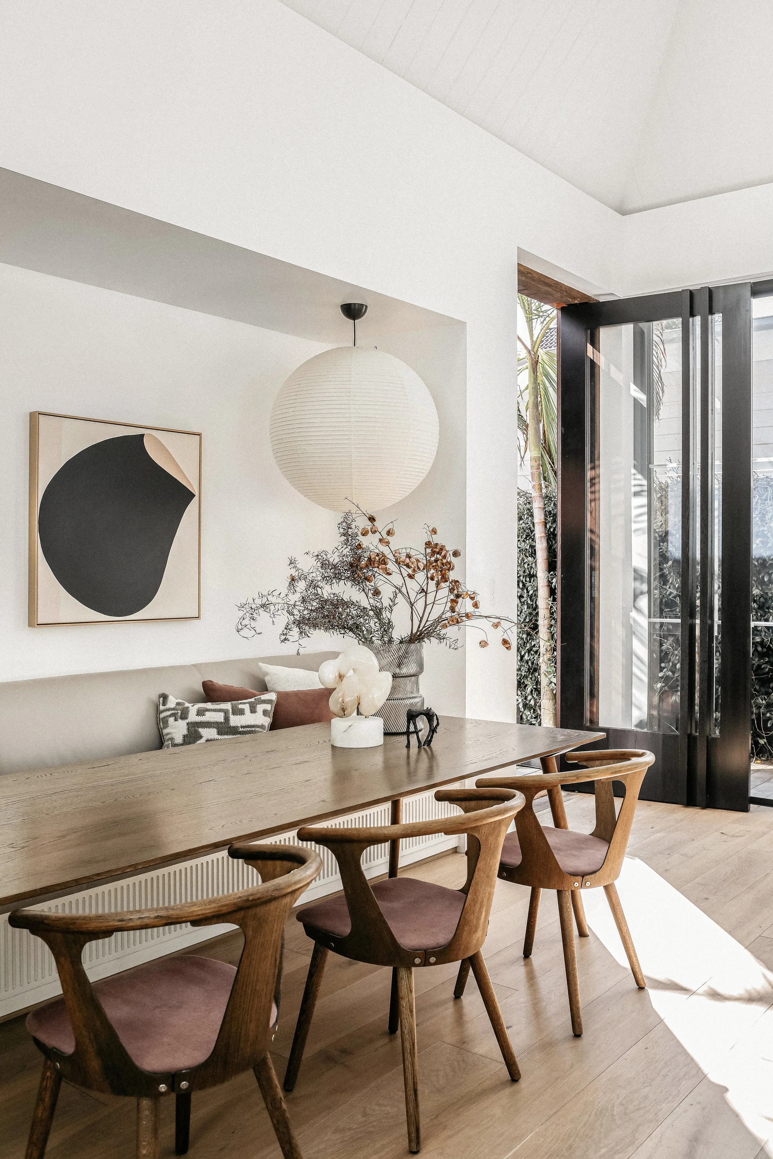 Modern dining room with a wooden table, four wooden chairs with mauve cushions, abstract art on a white wall, a paper lantern pendant light, a floral arrangement, and glass doors opening to outdoor greenery.