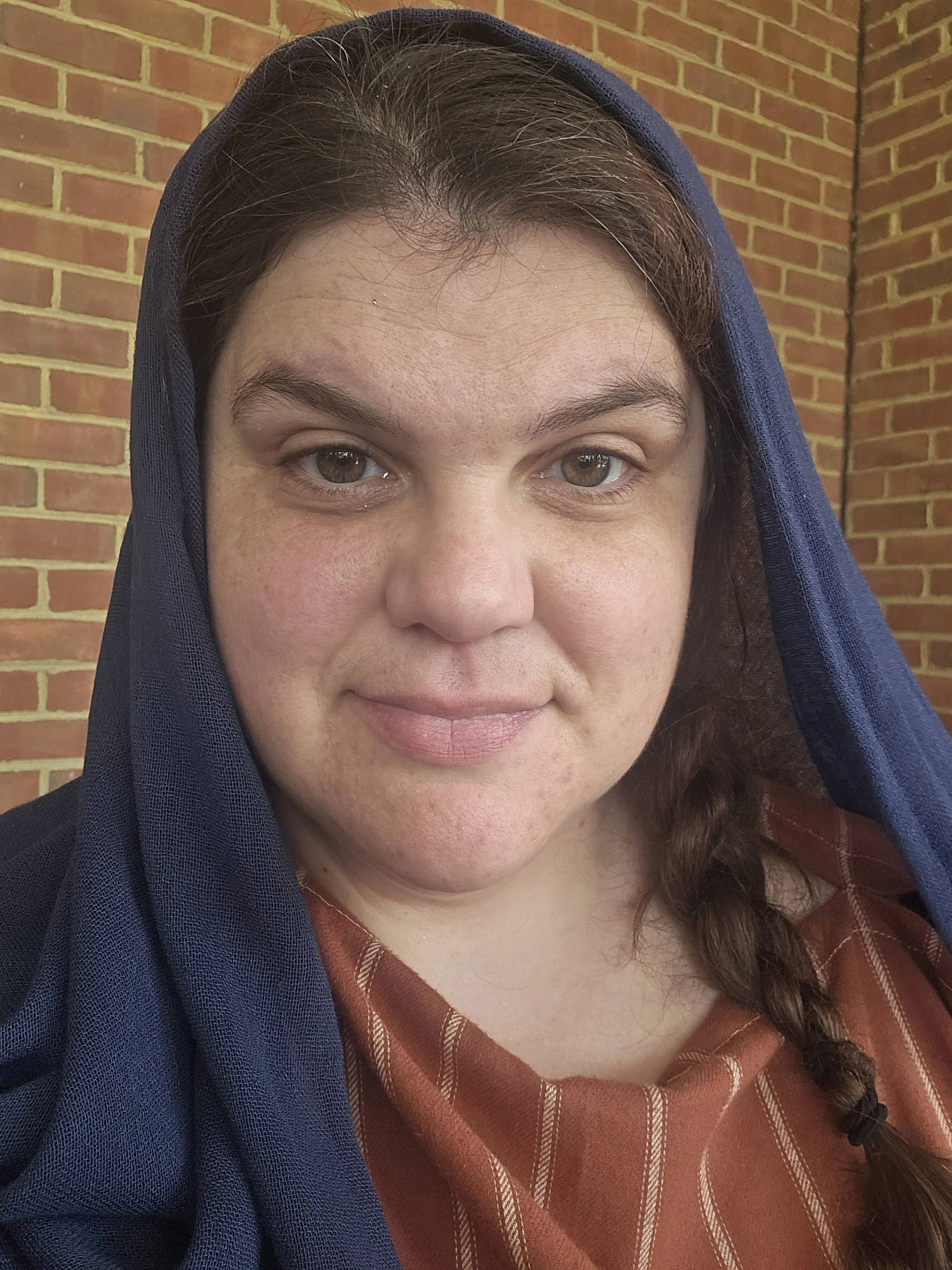 A woman with light skin and brown hair in a braid, wearing a blue scarf over her head and a brown top with white stripes, standing in front of a brick wall.