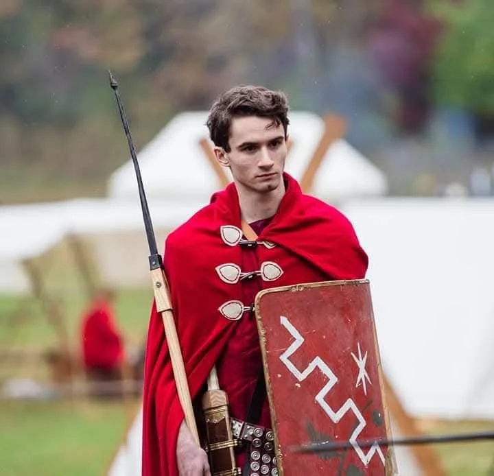 A young man dressed as a medieval warrior, wearing a red cloak, holding a spear and a shield with a white design, standing outdoors with tents in the background.