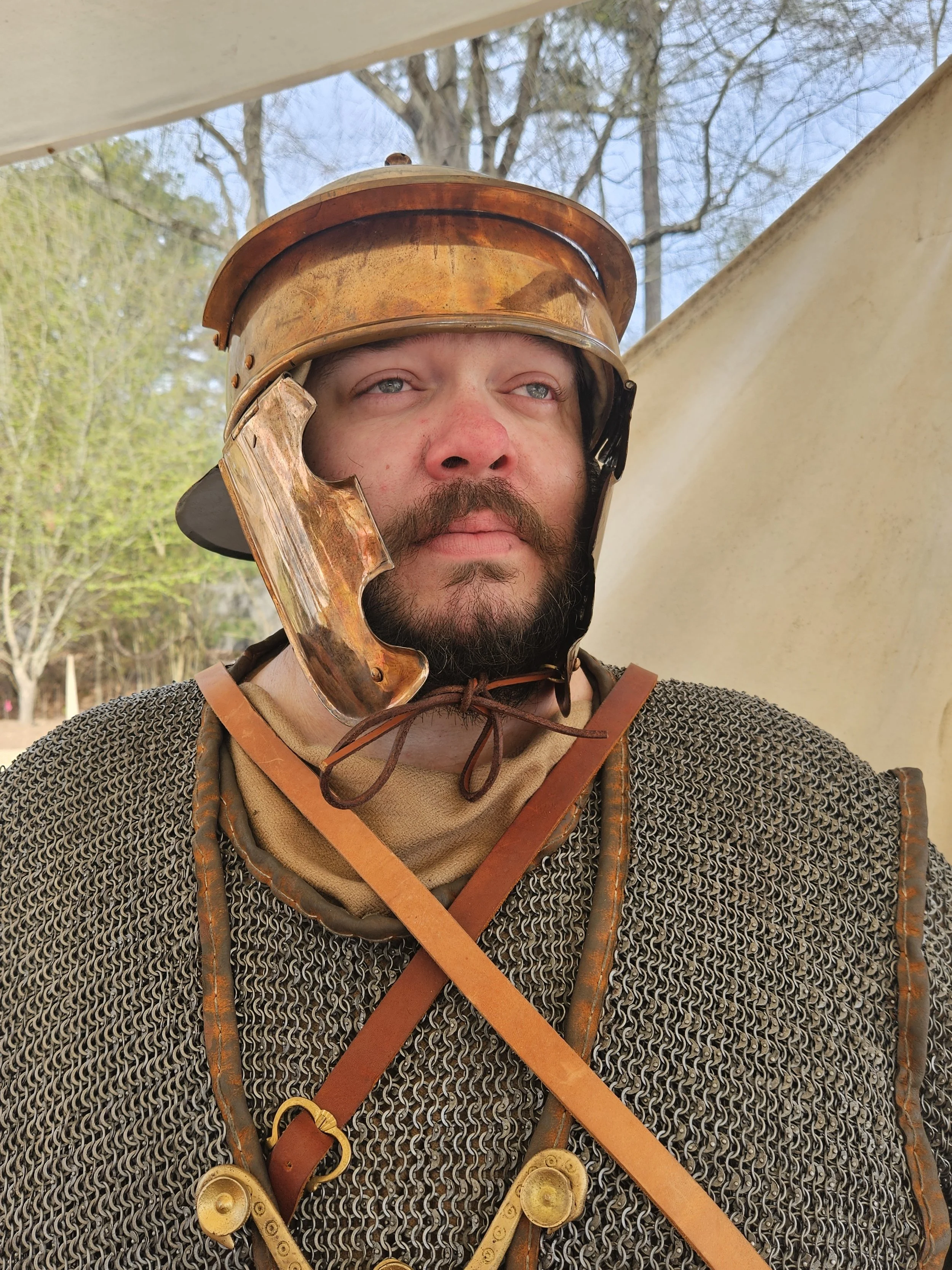 A man dressed as a medieval knight, wearing a chainmail shirt, leather straps, and a bronze helmet, standing outdoors with trees in the background.