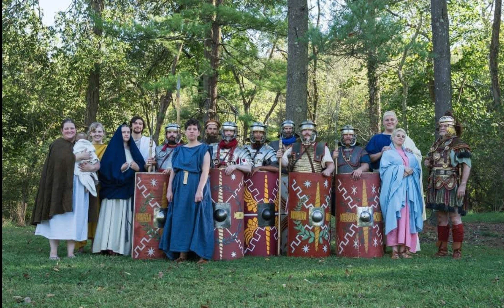 Group of people dressed in ancient Roman costumes, including soldiers with shields and helmets, standing outdoors in a wooded area.