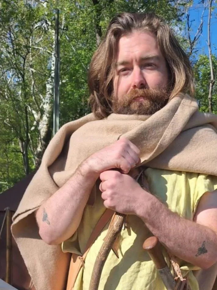 A man with long hair and a beard outdoors, wearing a beige shawl over his shoulders and holding a wooden staff, with green trees and a blue sky in the background.