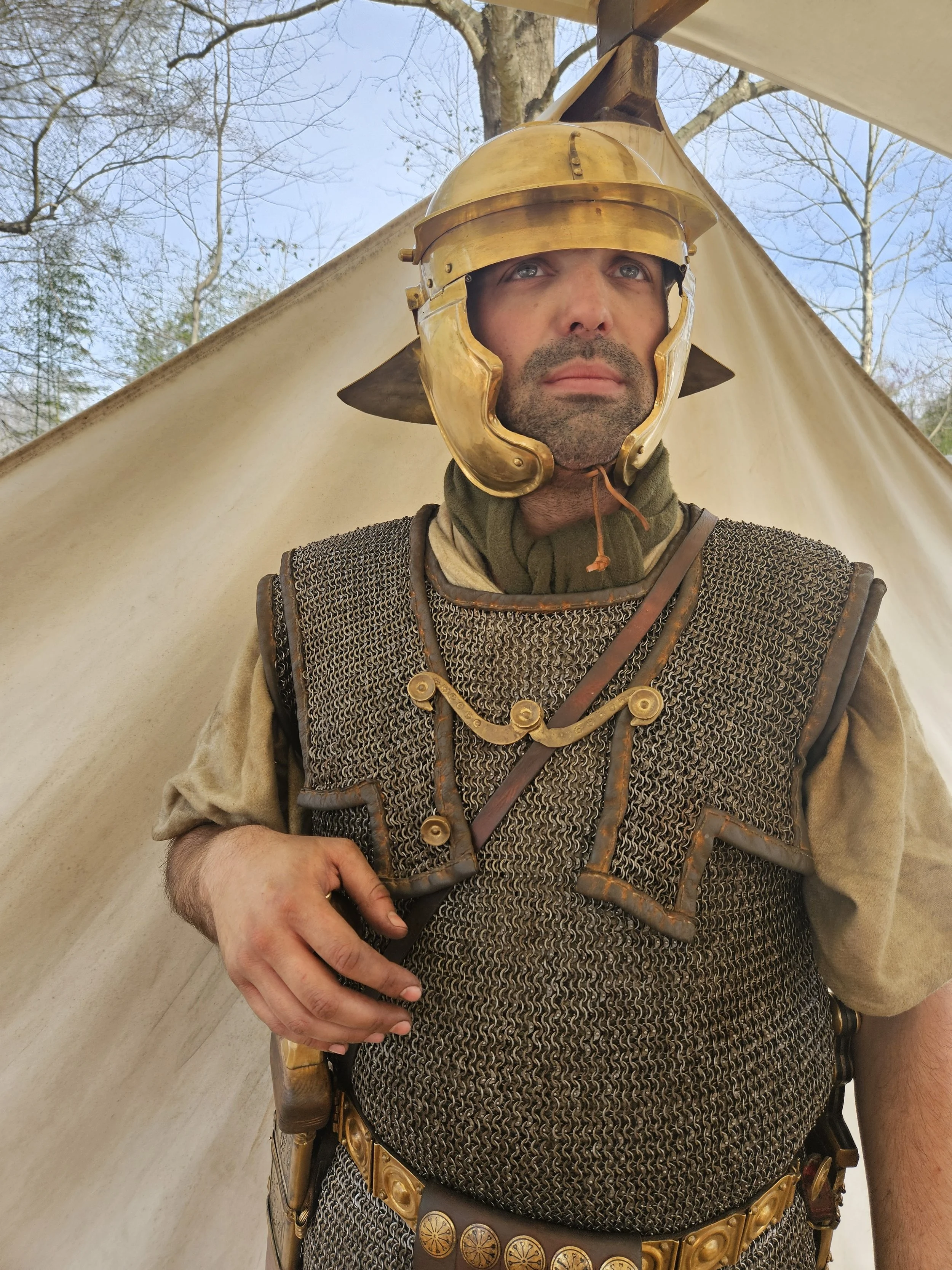 A man dressed as a Roman soldier wearing a gold helmet with a face guard, chainmail armor, and a decorative belt, standing outdoors with a tent and trees in the background.