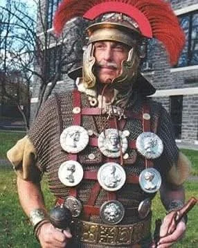 Person dressed as a Roman soldier in armor with medals and a red plume helmet, holding a small sword and a staff, outdoors with trees and a building in the background.