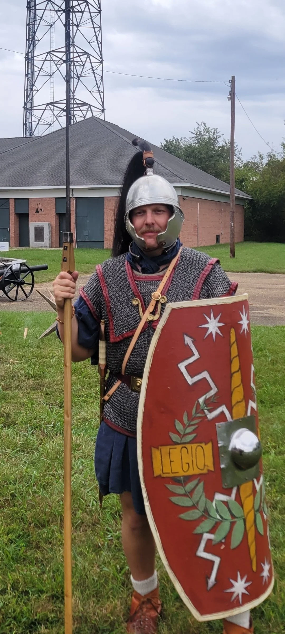 A man dressed as a Roman soldier, wearing a helmet, chainmail tunic, and shield with a sword and spear, standing outdoors on grass with a building, trees, and utility poles in the background.