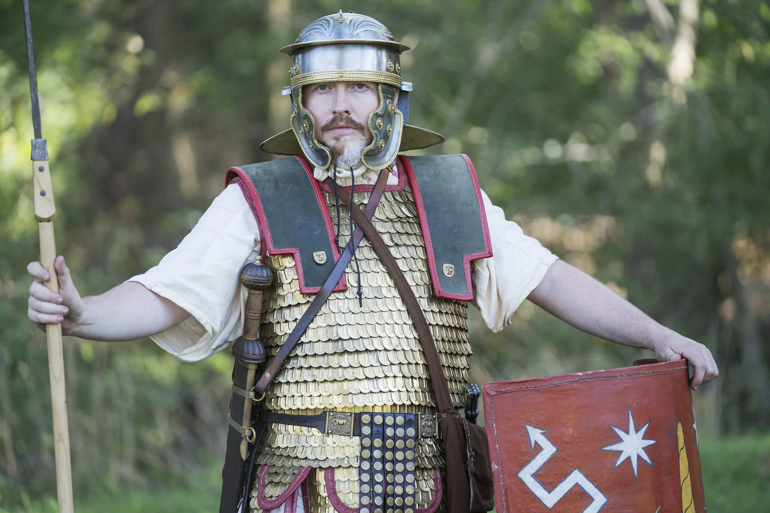 Person dressed as a Roman soldier holding a spear and shield, standing outdoors in a wooded area.