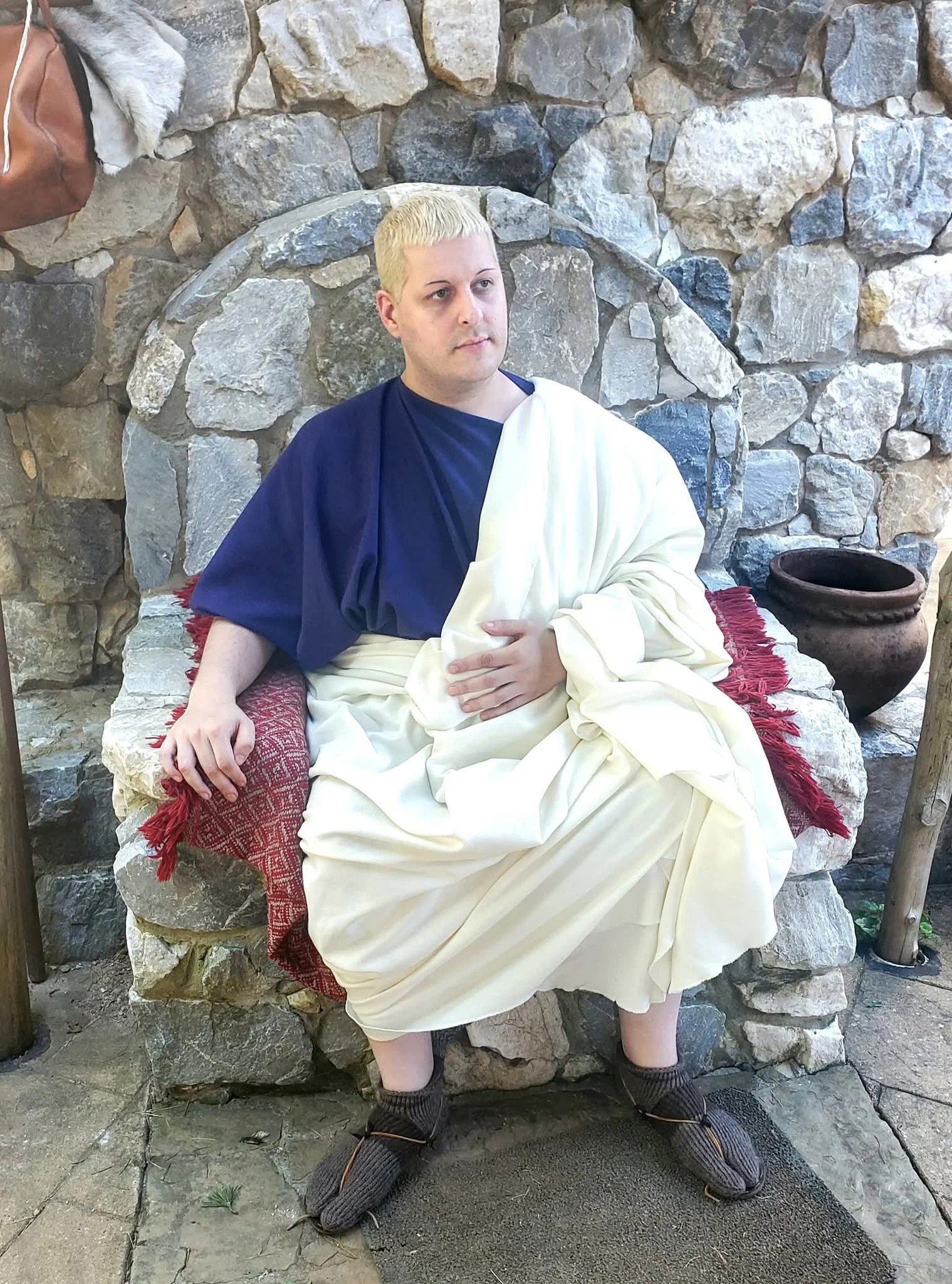 A person with short blond hair sitting on a stone bench dressed in a costume resembling ancient Roman attire, with a white toga and navy-blue shirt, in front of a stone wall.
